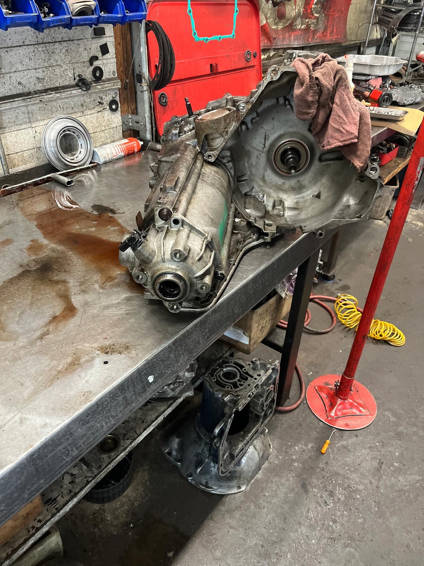 A dirty transmission on a workbench with oil stains, a red toolbox in the background, and shop tools.