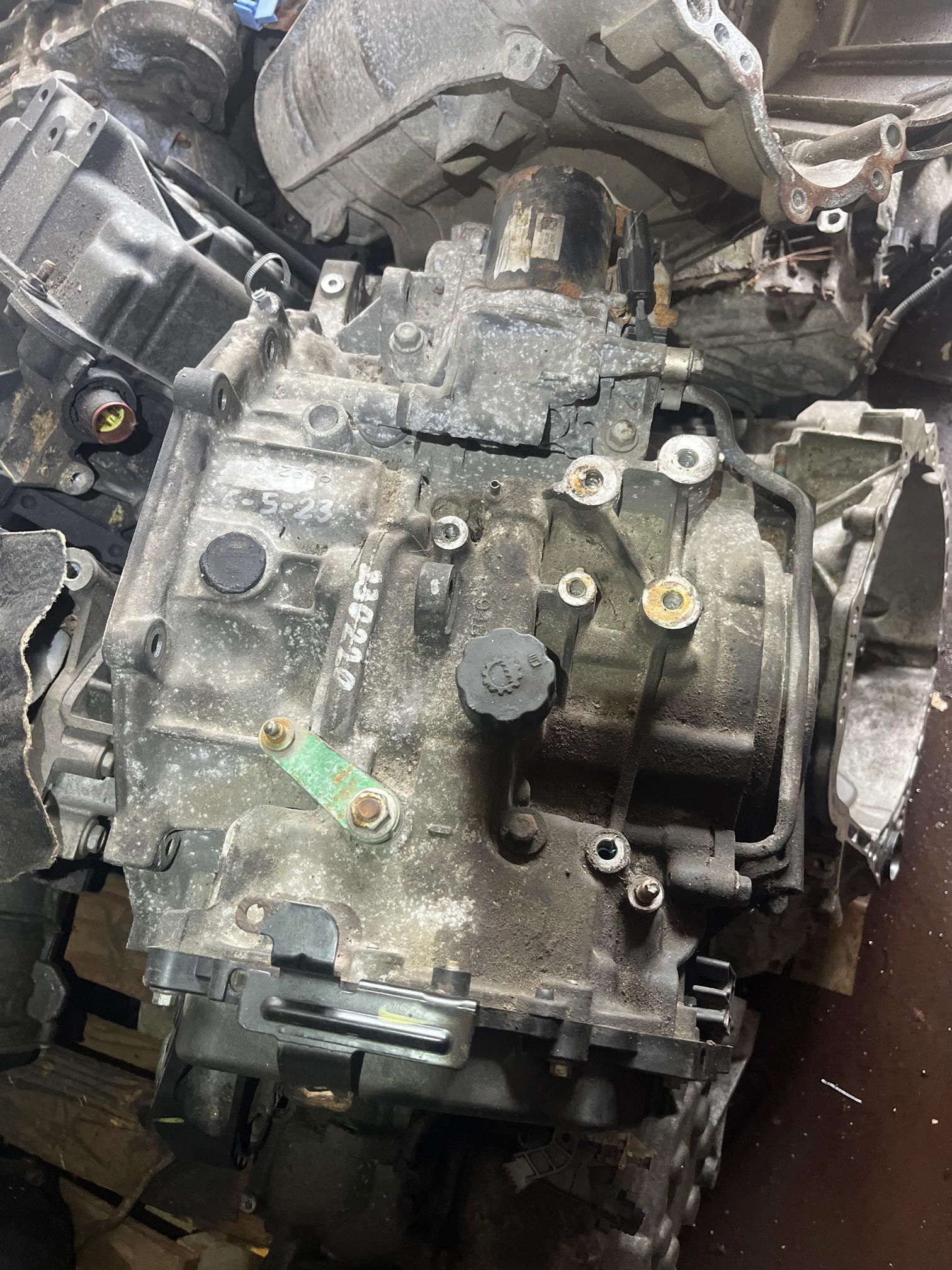 A used, dirty car transmission in a pile with others. The unit has various nuts and bolts and is metallic.