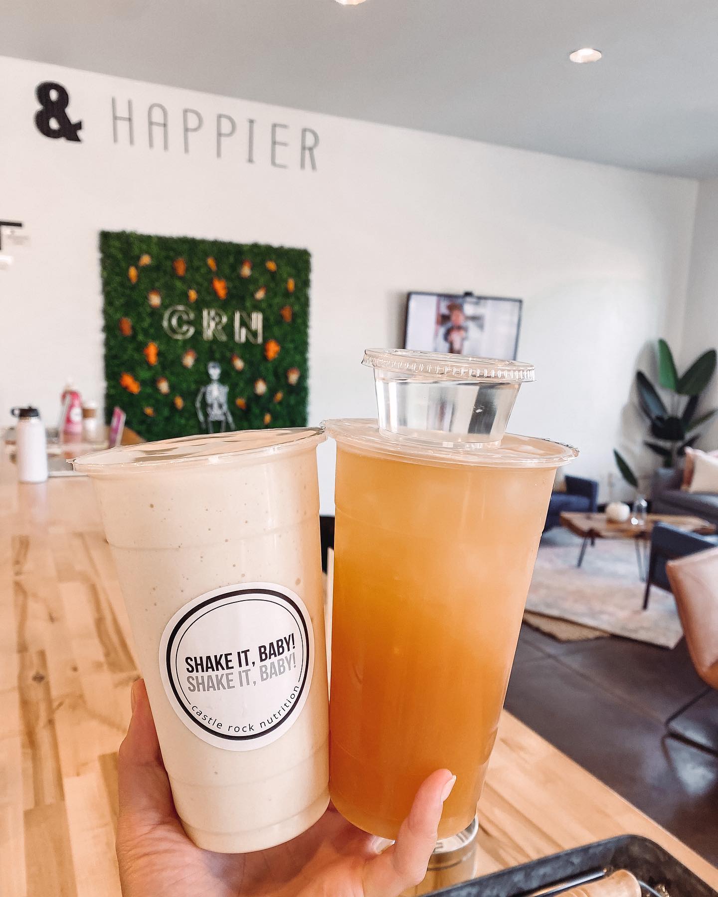A person is holding two cups of drinks in front of a sign that says & happier
