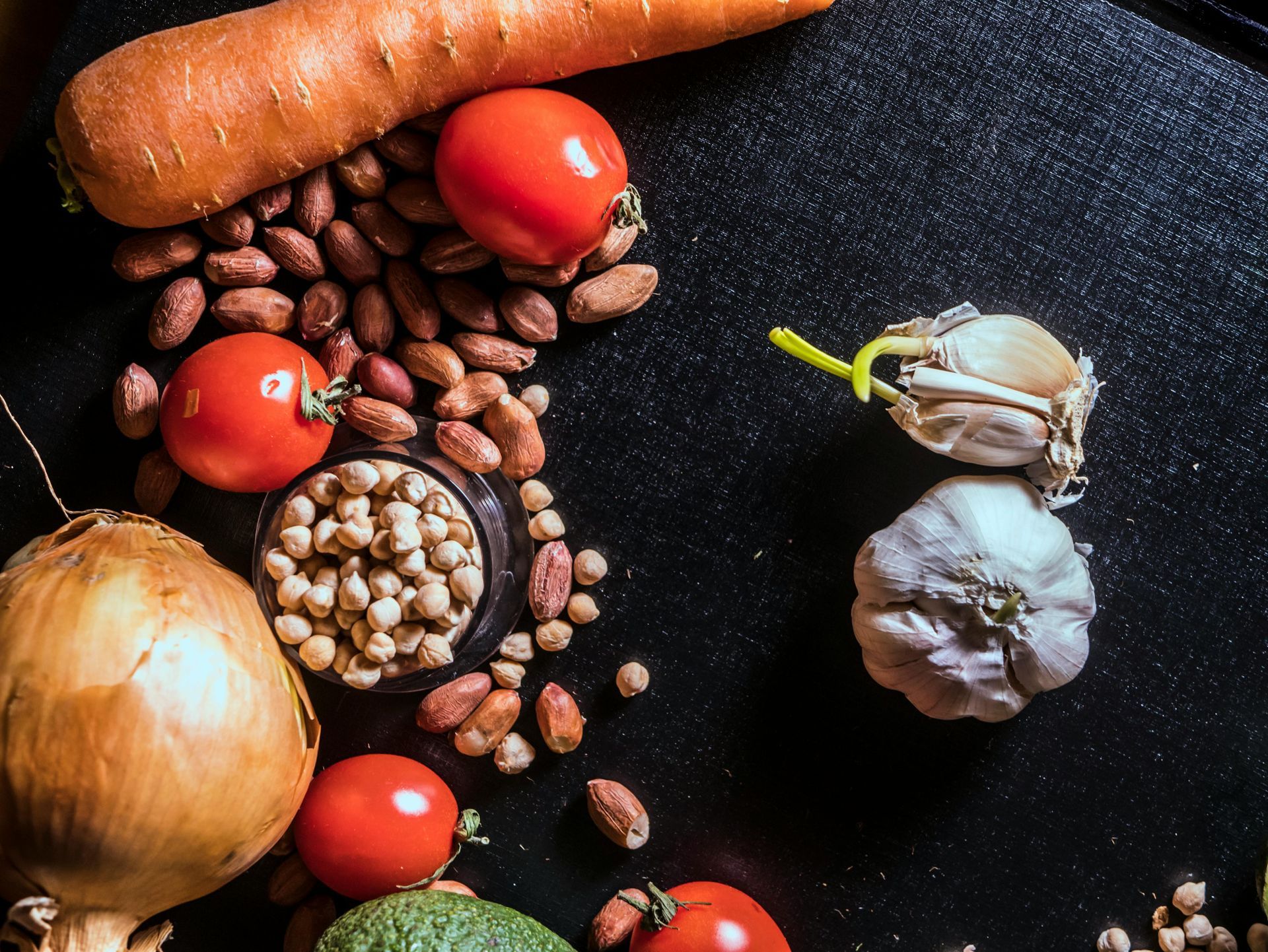 Fresh vegetables and legumes arranged on a dark surface: onion, carrots, tomatoes, garlic, and beans.