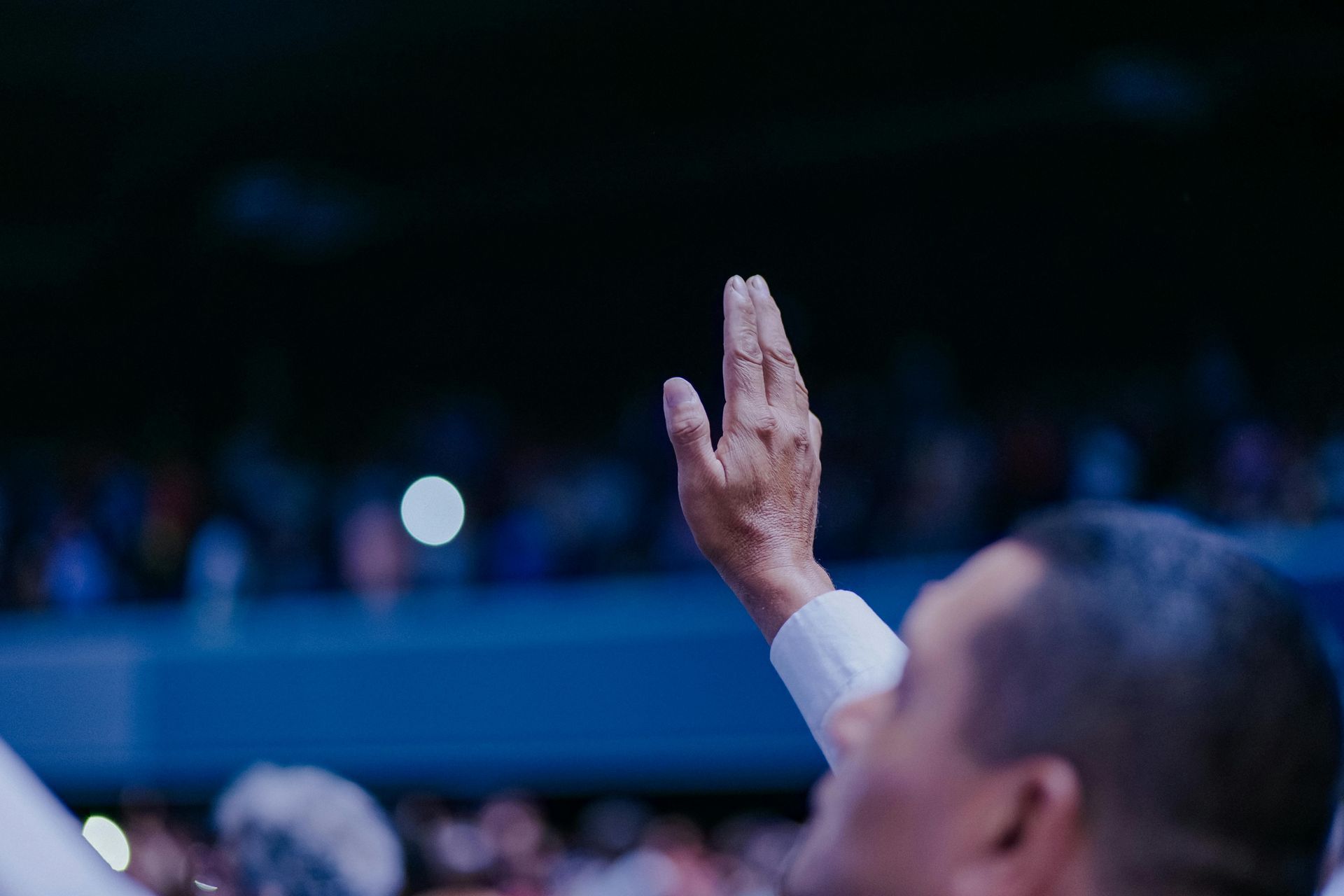 Person raising a hand in a dimly lit auditorium, with a blurred audience in the background.