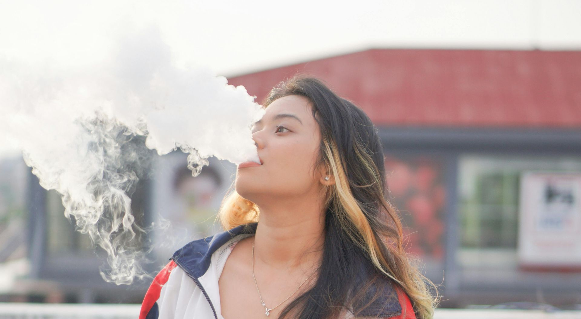 Woman exhaling smoke outdoors, wearing a white and red jacket with blurred buildings in the background