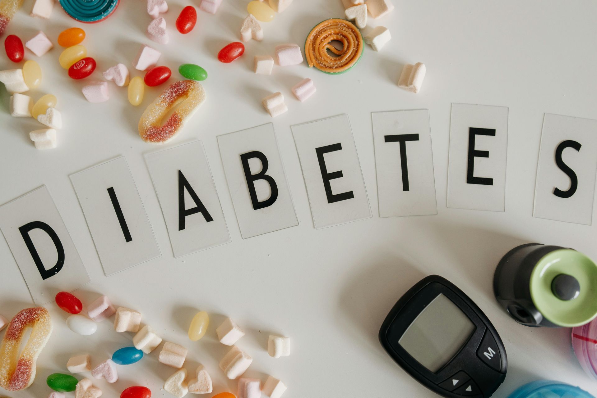 Diabetes spelled with tiles, surrounded by sweets and a glucose meter on a white surface