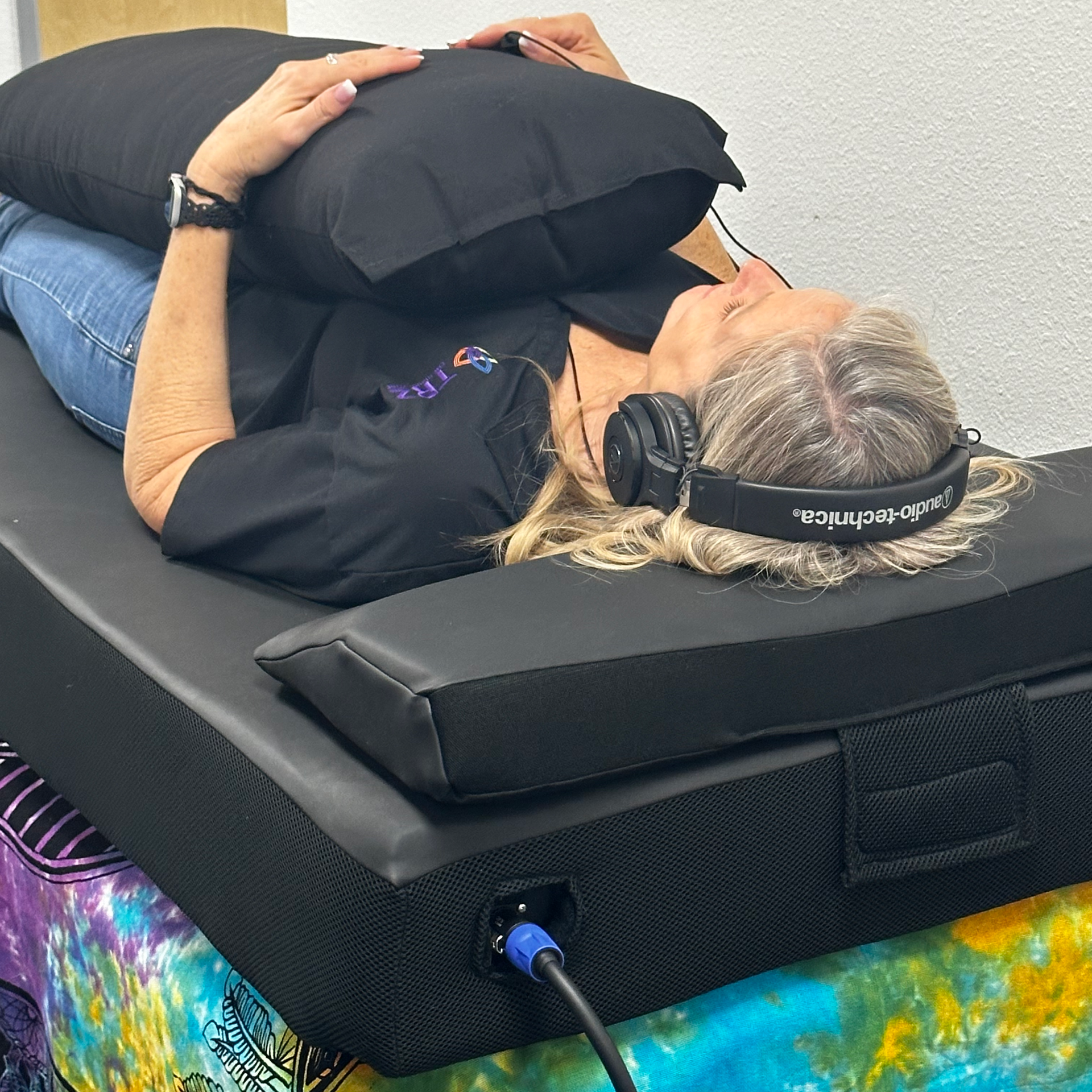 A woman is laying on a bed wearing headphones and holding a black pillow.