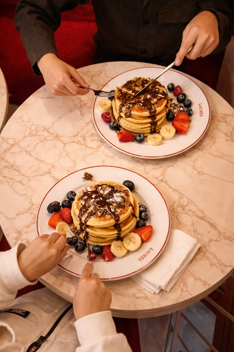 Due persone sedute a un tavolo di marmo mangiano pile di pancake guarniti con sciroppo al cioccolato, fragole e mirtilli.