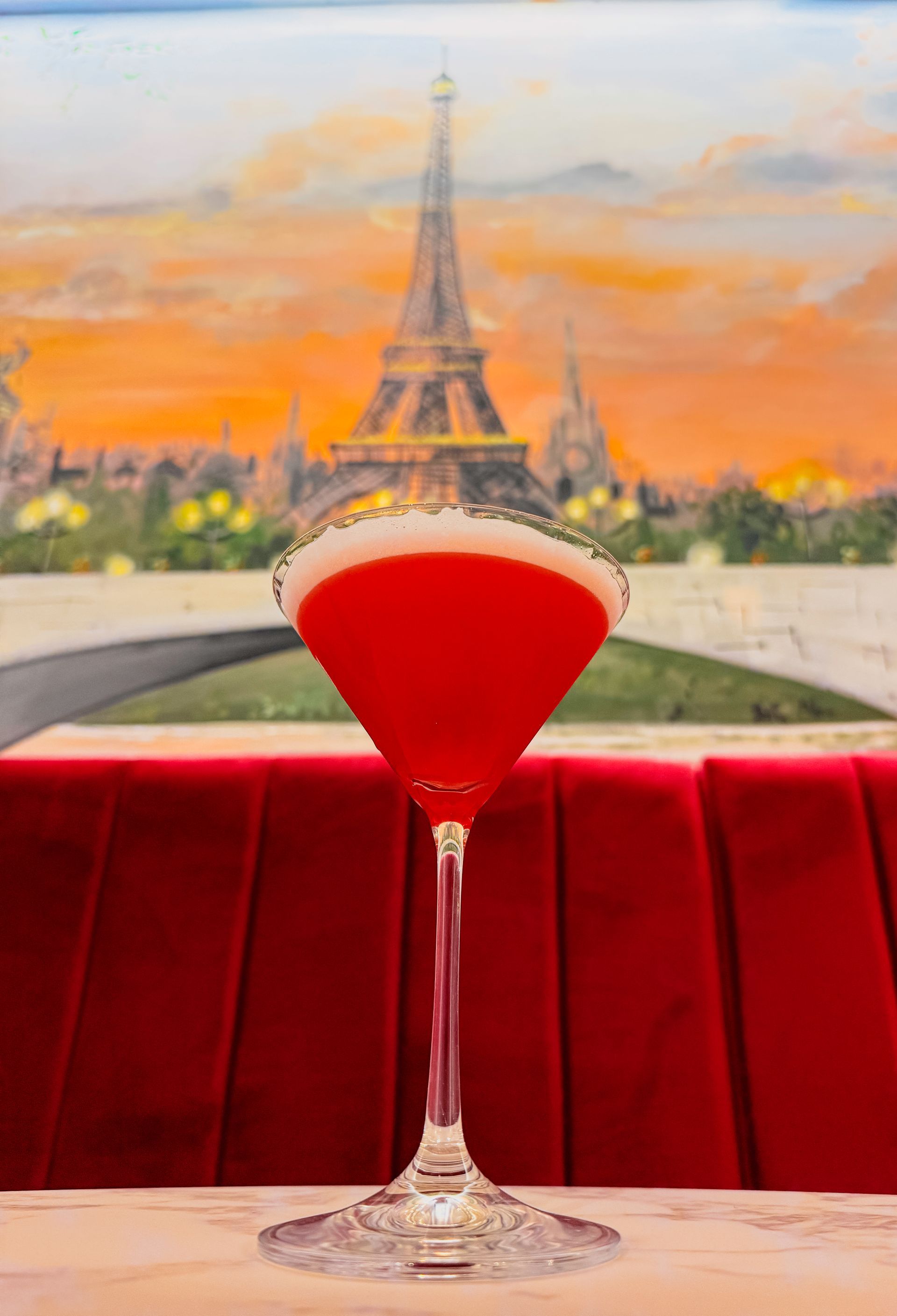 Un cocktail rosso in un bicchiere con stelo è appoggiato su un tavolo di fronte a un murale raffigurante la Torre Eiffel al tramonto.