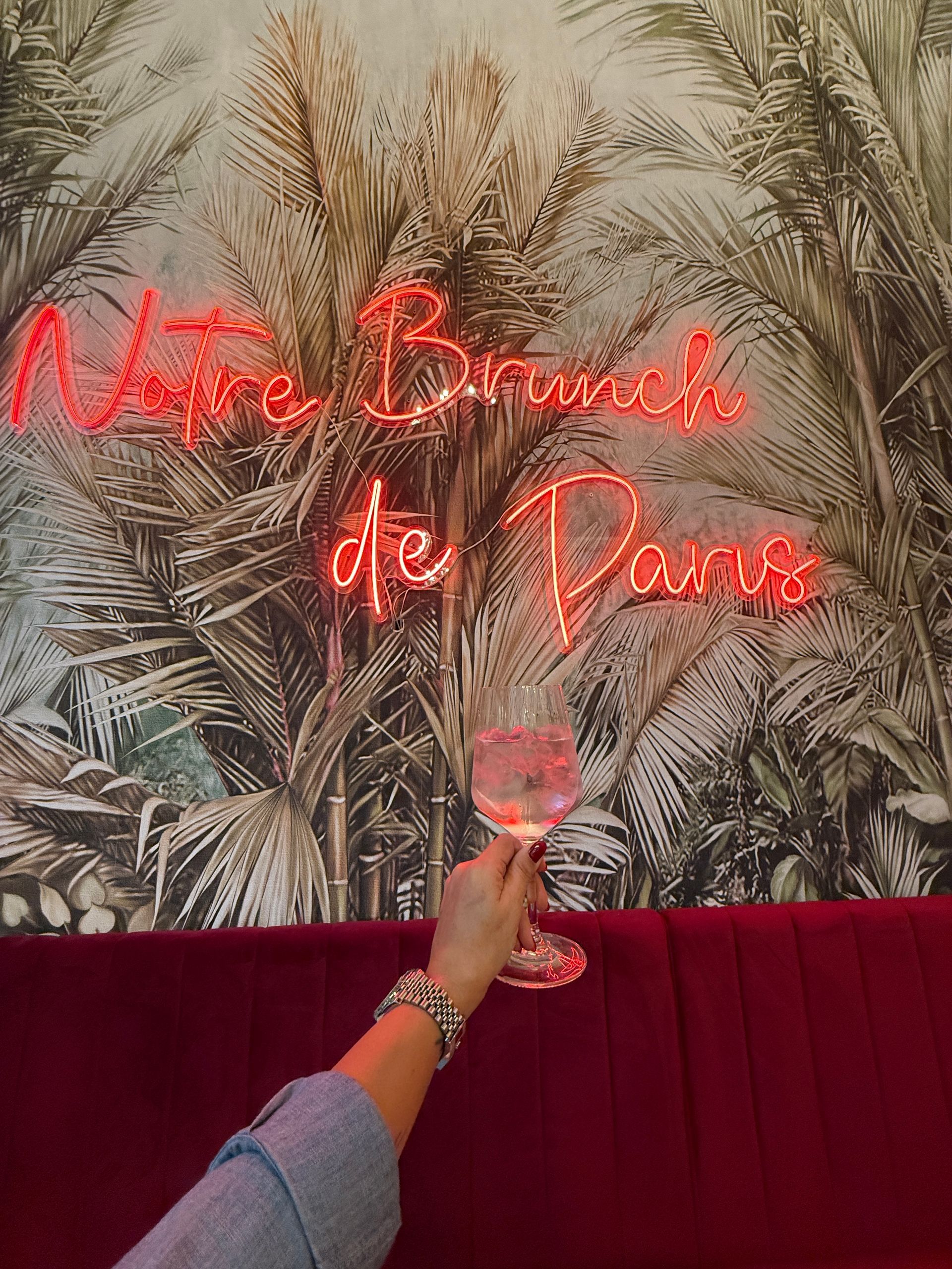 Una mano regge un bicchiere di bevanda rosa sullo sfondo di una carta da parati con foglie di palma e un'insegna al neon con la scritta 