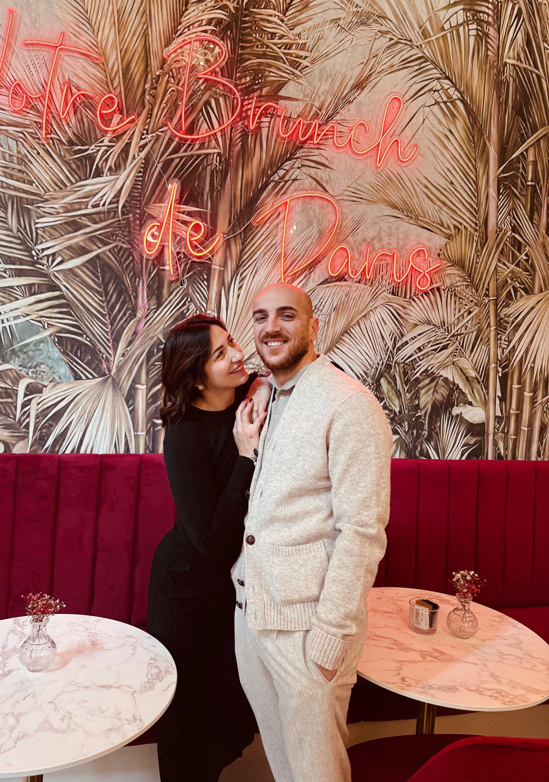 Una coppia in piedi, a braccetto, davanti a un murale raffigurante foglie di palma con scritte al neon rosse e divanetti rosa scuro.