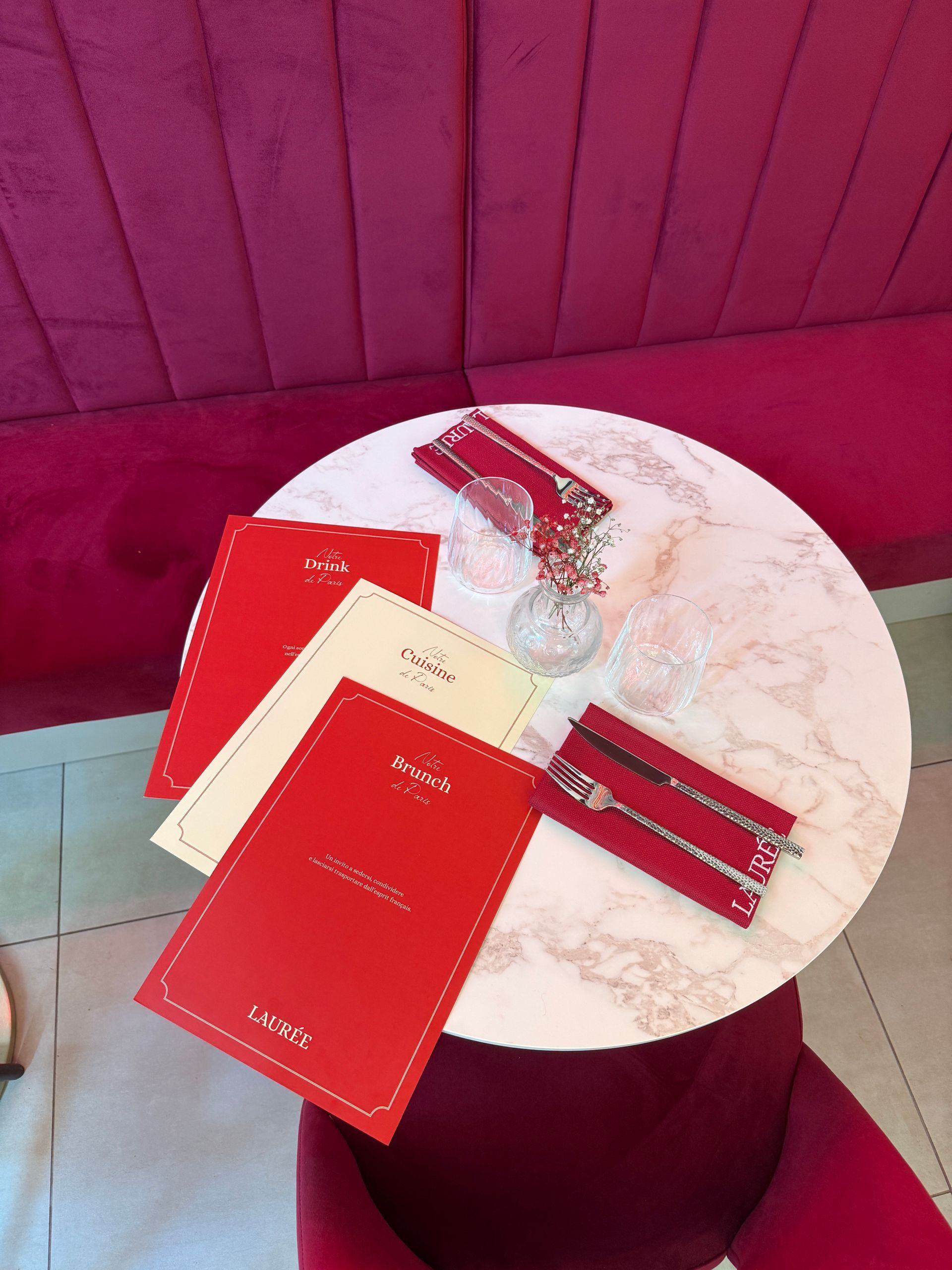 Un tavolino da caffè con piano in marmo, completo di menù, posate e bicchieri, posizionato di fronte a una panca imbottita di colore rosa intenso.