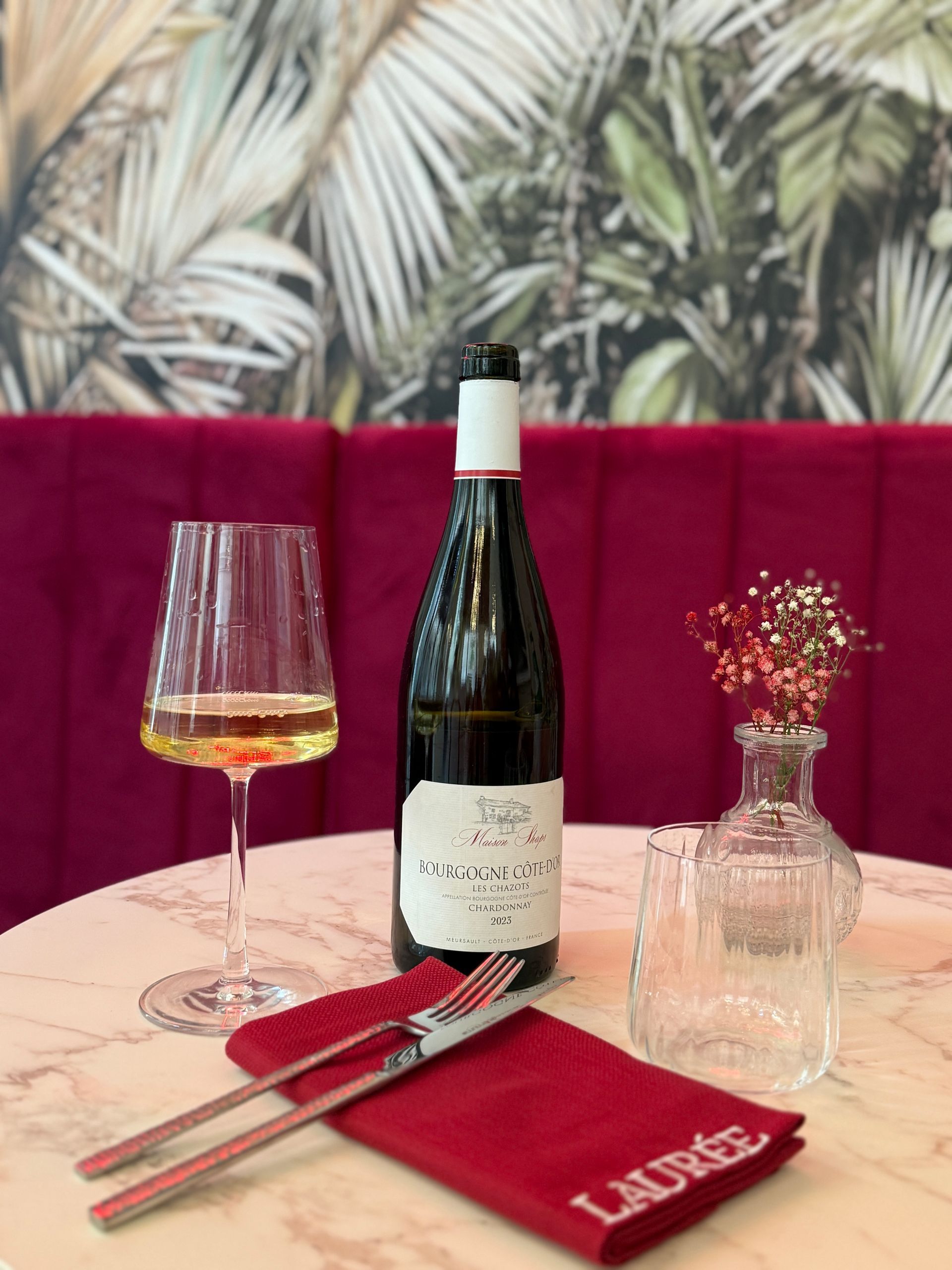 Una bottiglia di vino e un bicchiere pieno sono appoggiati su un tavolo di marmo con un tovagliolo color bordeaux, un vaso trasparente e una carta da parati a motivi botanici.