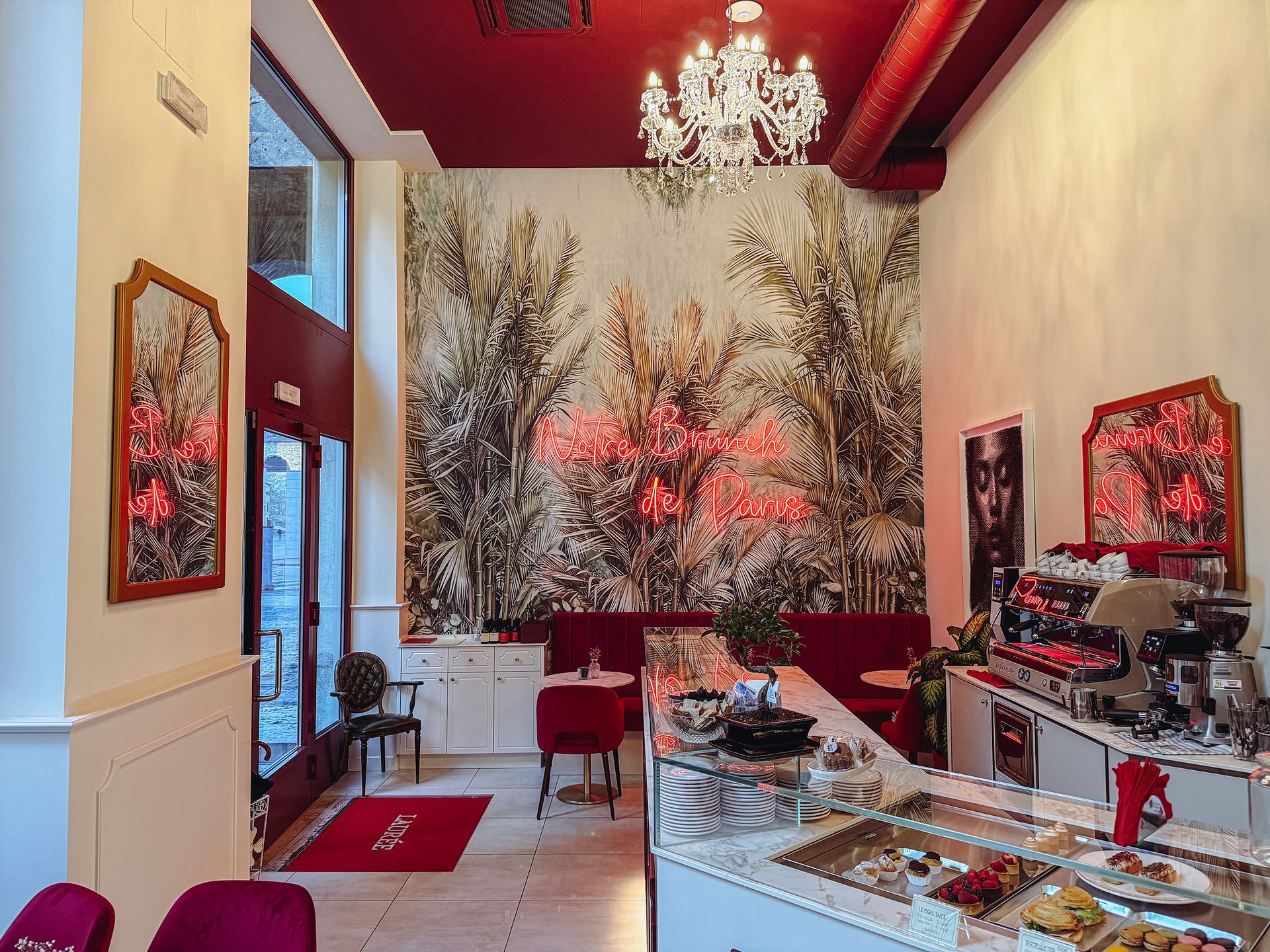 Vista interna di una caffetteria con soffitto rosso, carta da parati decorativa, lampadario di cristallo e bancone di servizio in marmo.