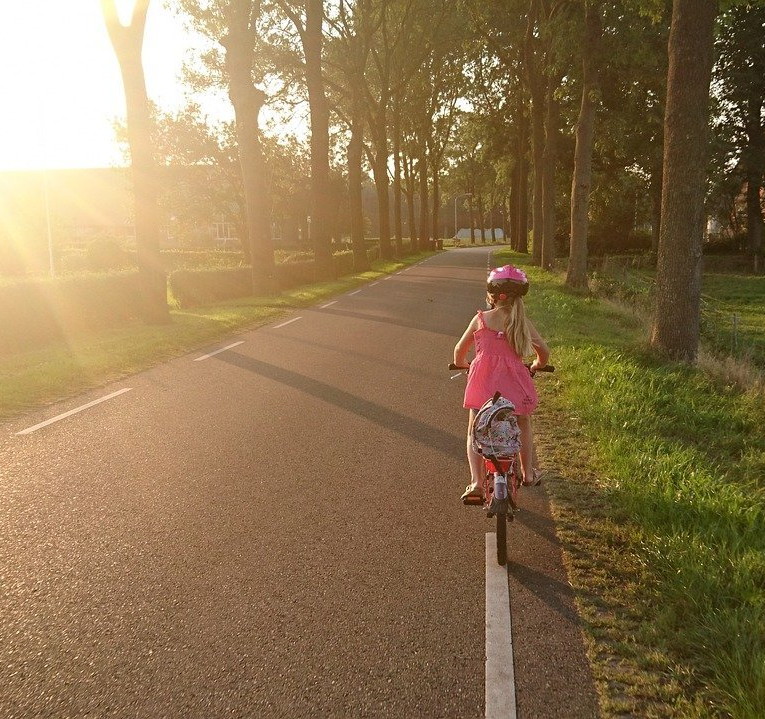 The first image is of a young girl biking down an asphalt road at dusk. The second picture is of a mother and father walking across a crosswalk in a busy intersection. The father has the baby on his shoulders.