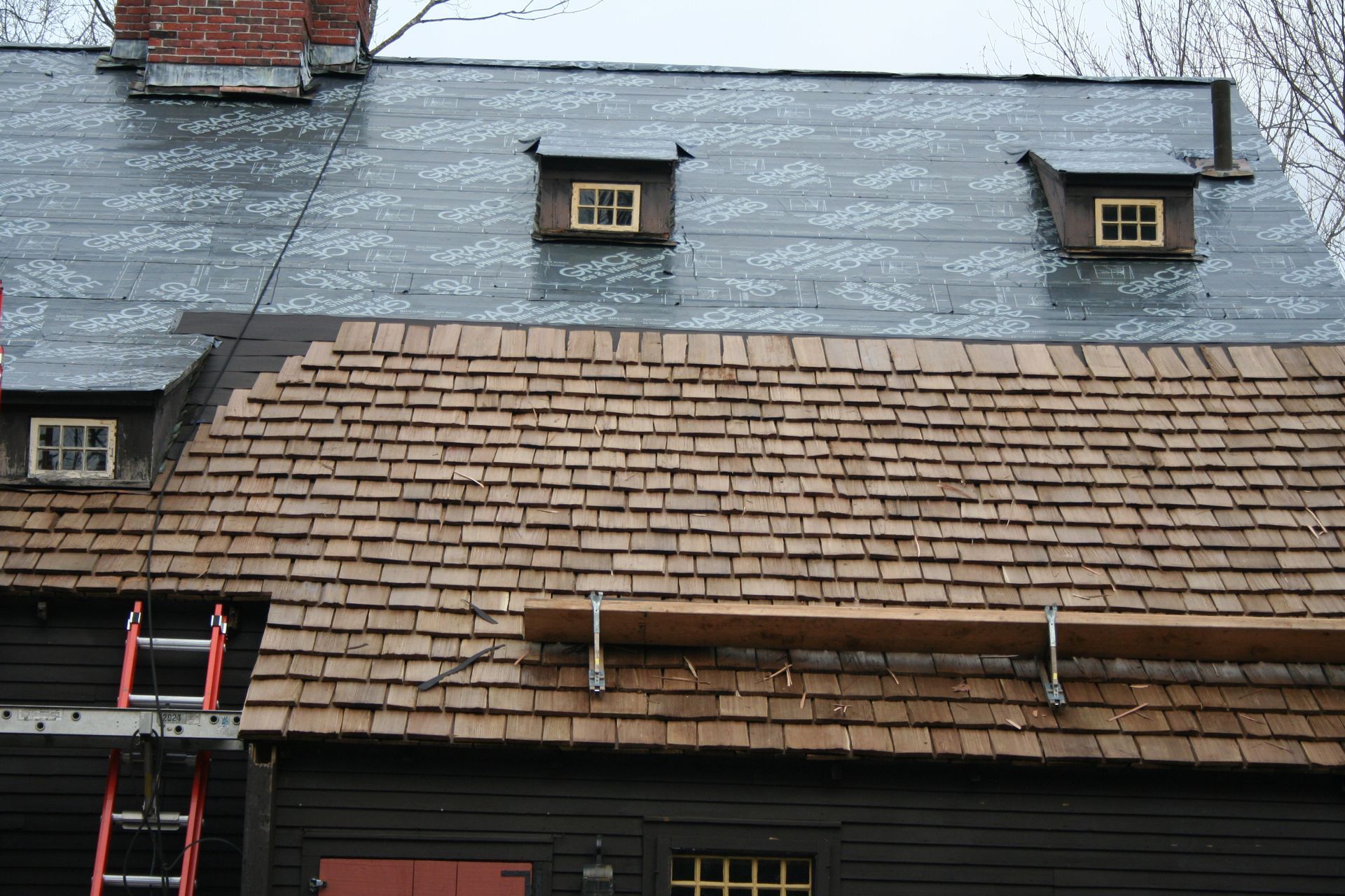 A partially shingled roof on a dark building with three dormer windows and a ladder.