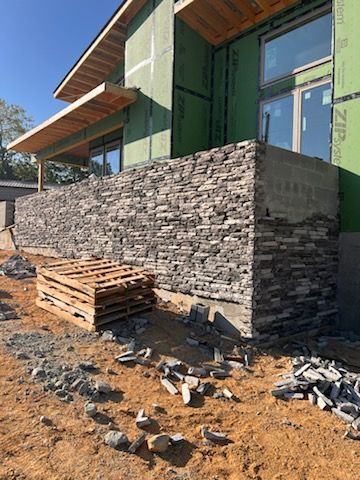 Exterior house under construction; stone veneer wall, wooden beams, pallets, and dirt ground.