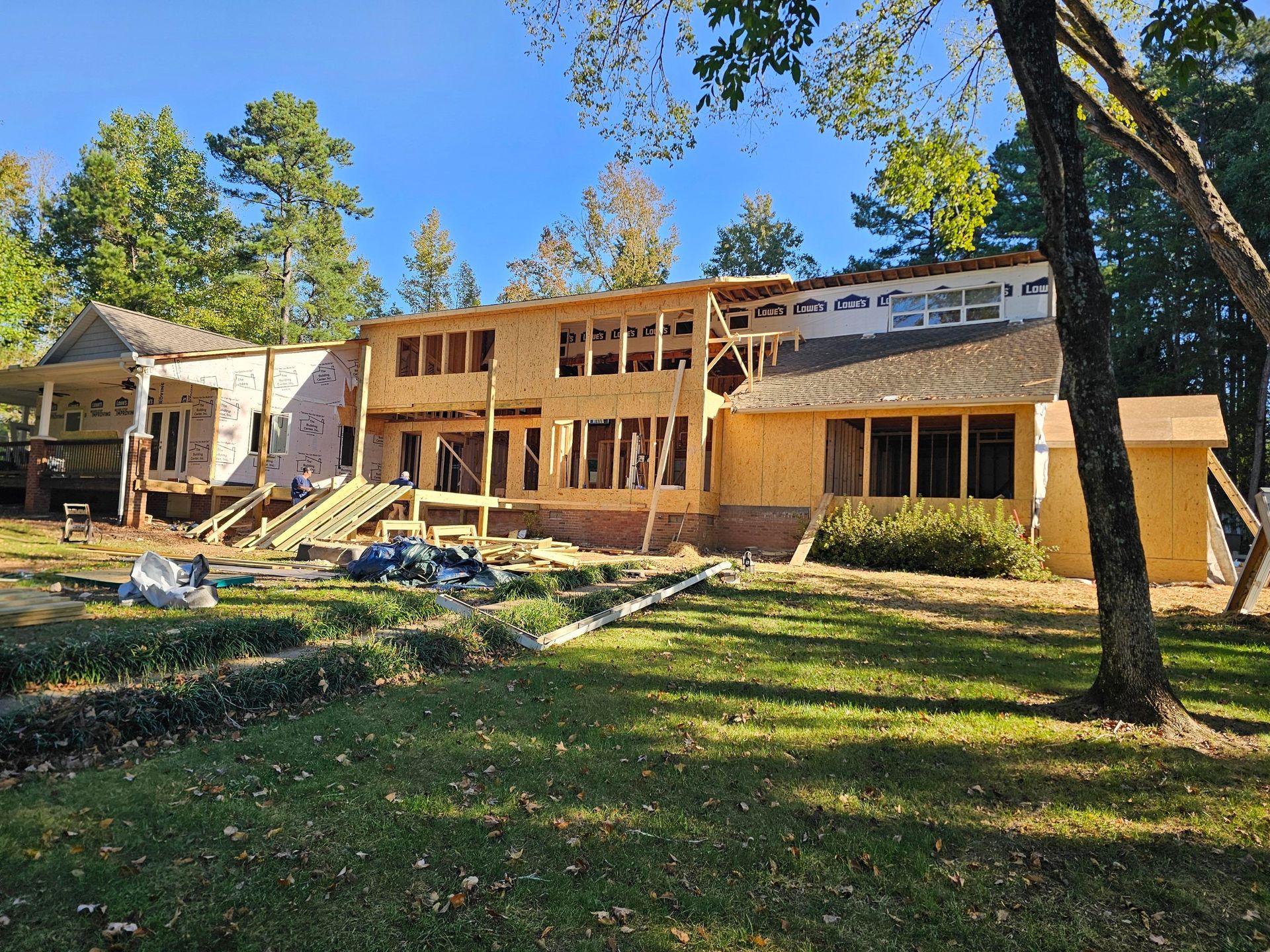 House under construction; exposed wood framing, sheathing; blue tarp; green grass; trees in background.