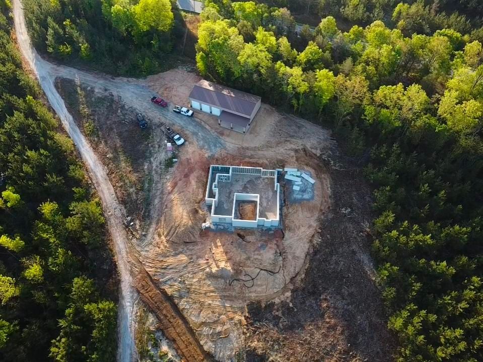 Aerial view of a construction site. Concrete foundation, surrounding earth clearing, shed, vehicles, and a dirt road in a wooded area.