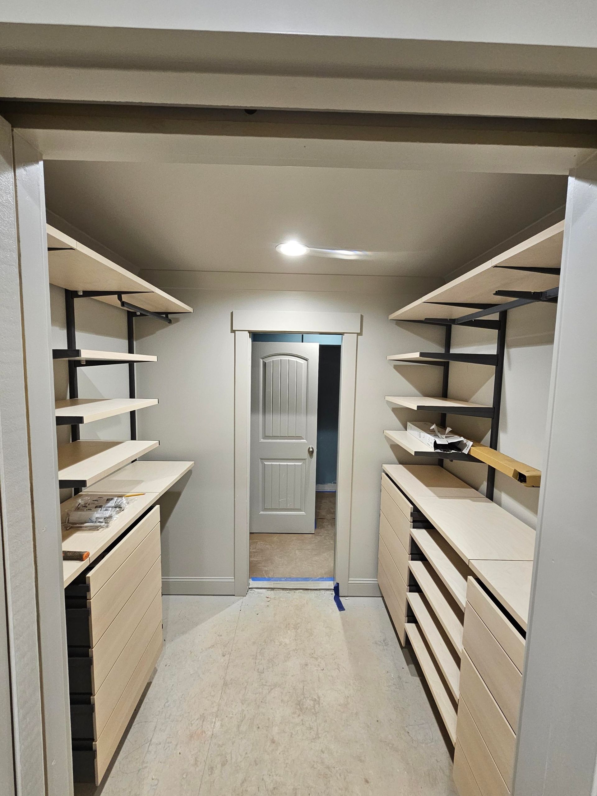 Walk-in closet with shelves and drawers on both sides, a door at the end. Beige wood and gray walls.