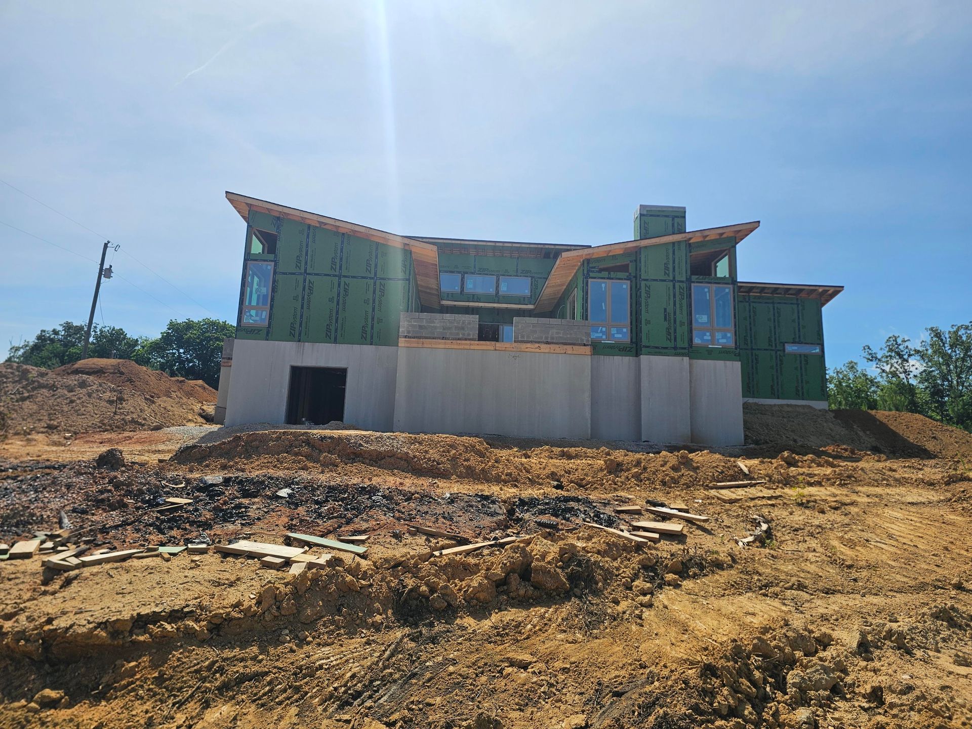 A house under construction with exposed framing and large windows, on a dirt lot under a bright sky.
