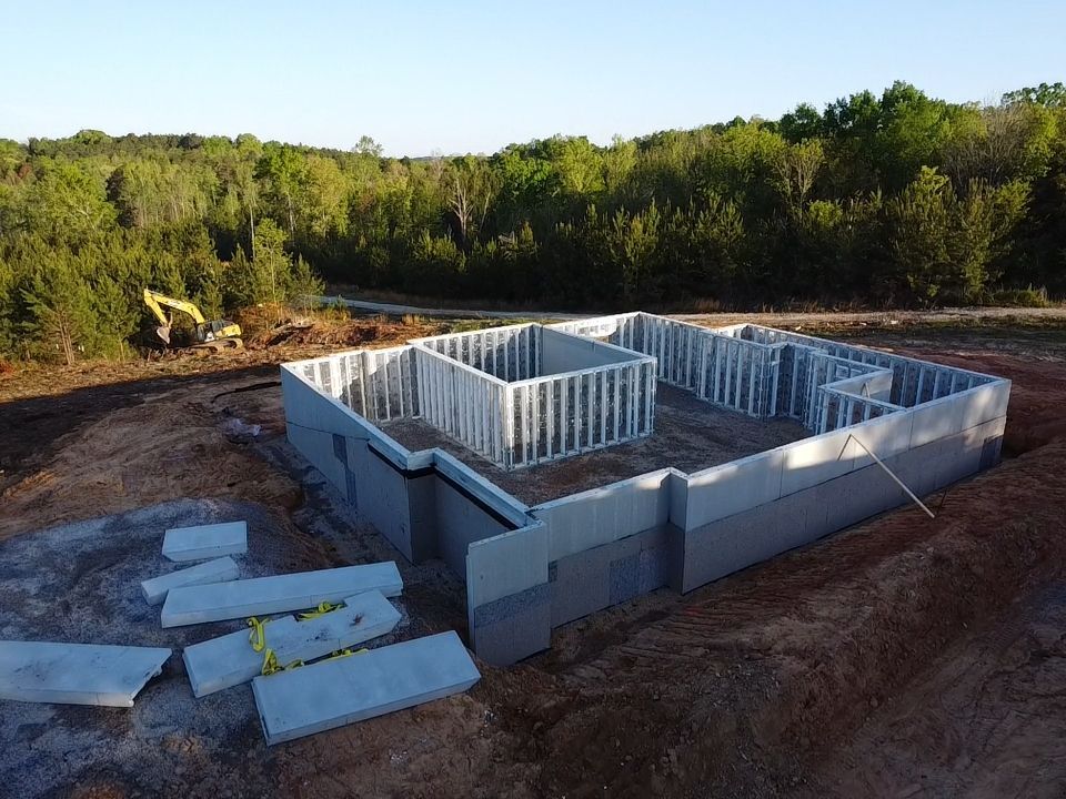 Foundation of a building under construction, grey concrete walls, metal framing, dirt, yellow safety straps, trees in the background.