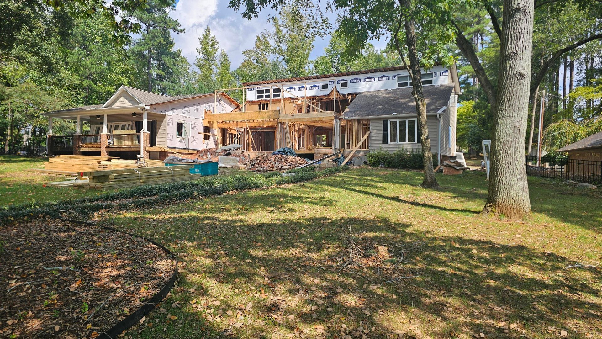 House under construction with exposed framing. Lumber piles, trees, and blue sky visible.