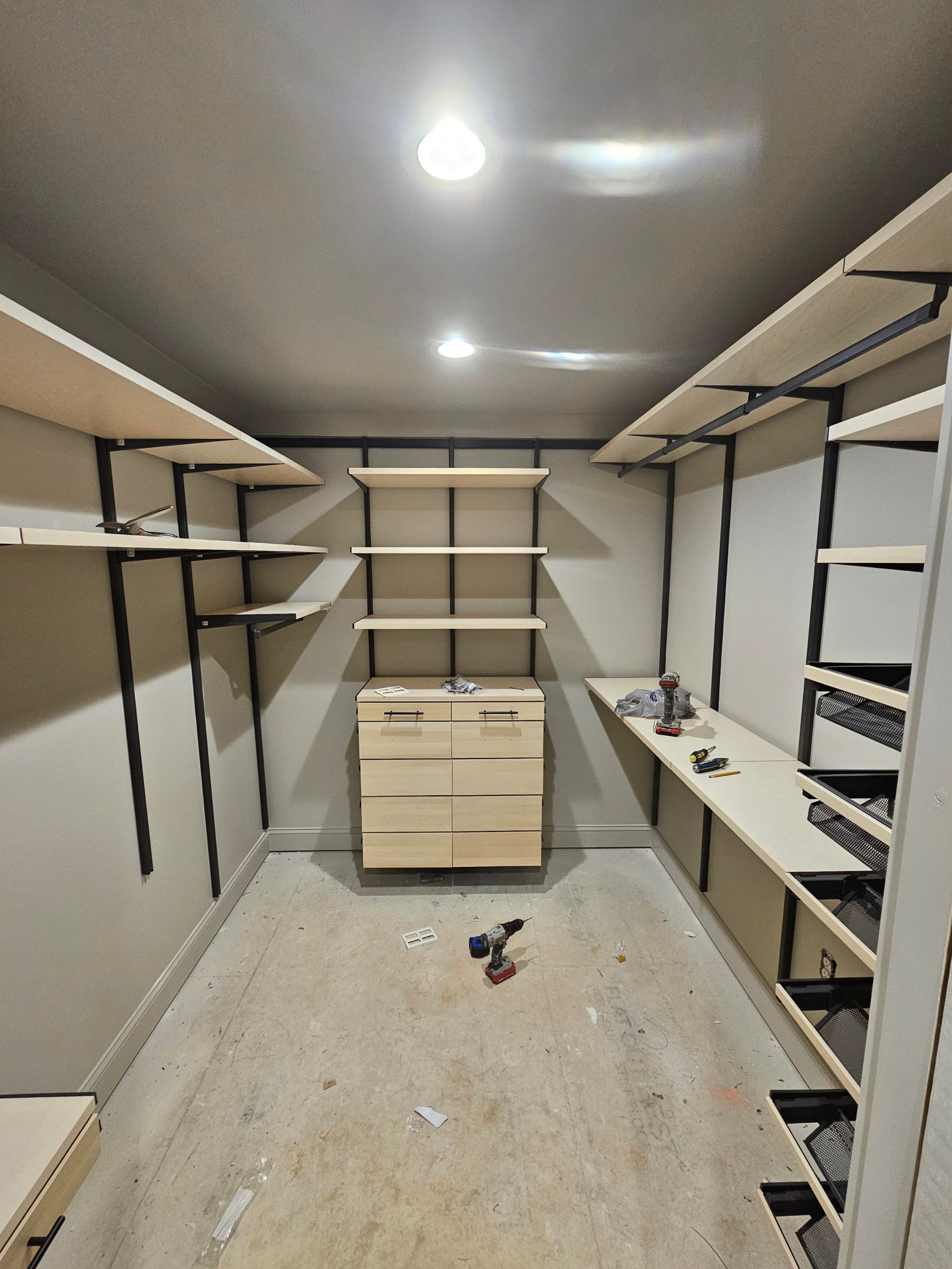 Empty walk-in closet under construction with shelves and drawers. Light wood and black metal supports against neutral walls.
