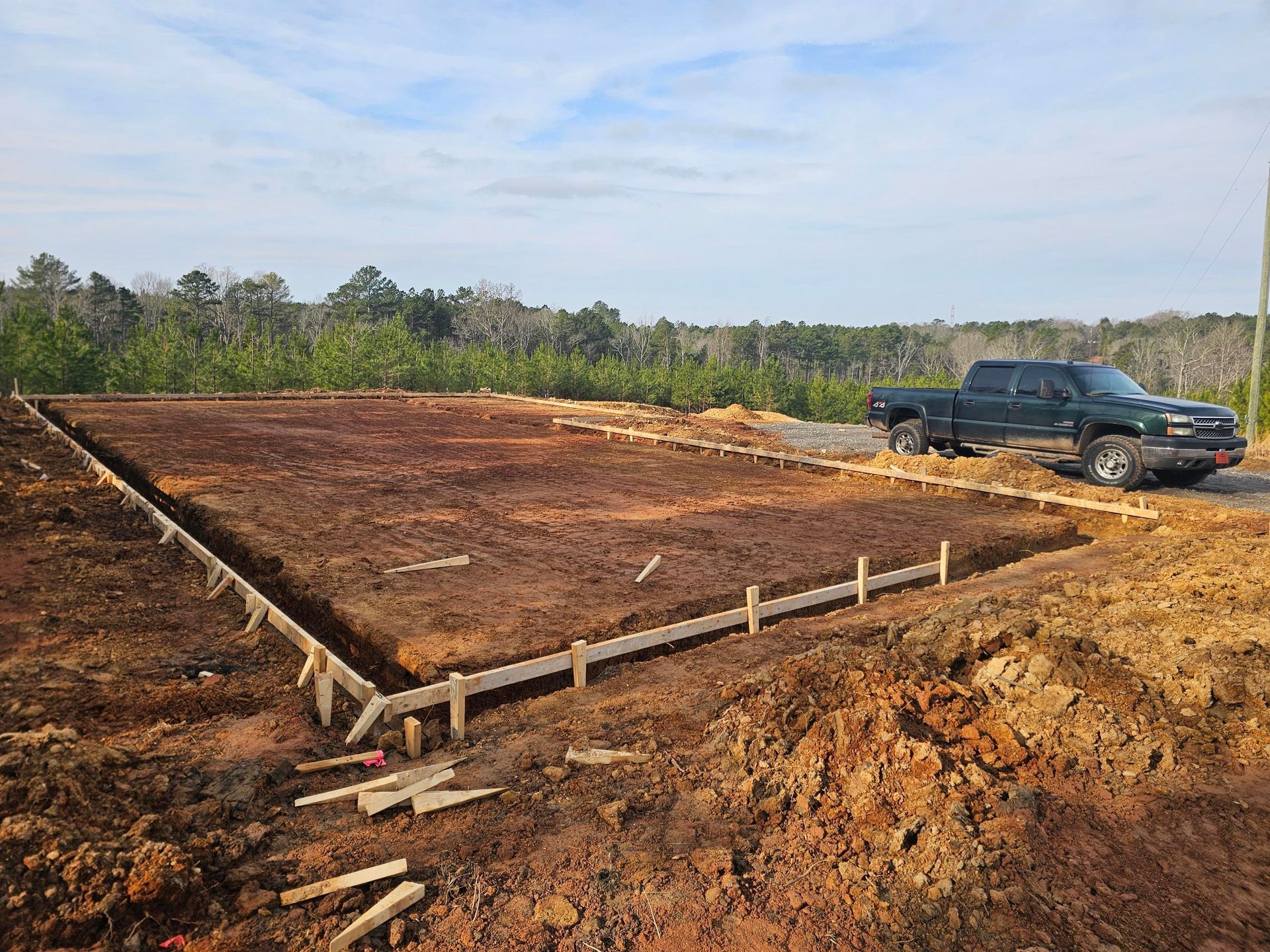 Foundation frame on dirt ground with a truck parked nearby, against a backdrop of trees and sky.