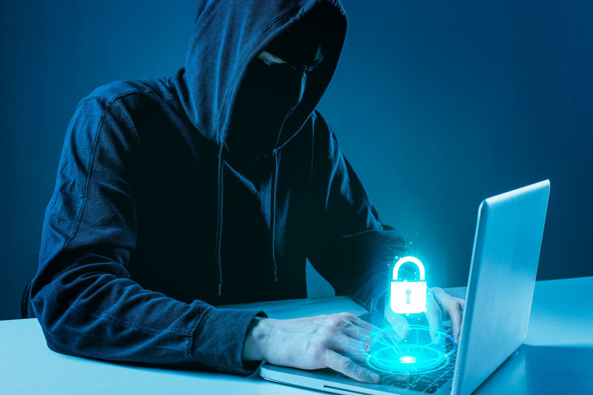 Person in hoodie and mask working on a laptop, with a glowing padlock graphic superimposed on the screen.
