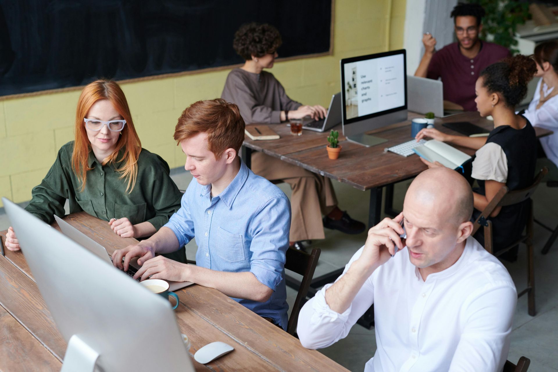 People working on computers at a wooden table in an office. One person is on a call.