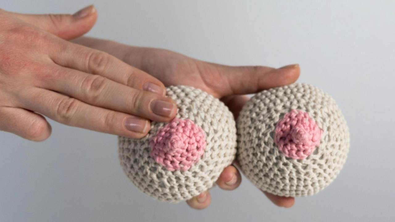 A hand touching two crocheted breast models, beige with pink areolas.