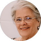 Mulher sorridente com cabelos grisalhos e óculos; colar de pérolas.