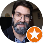 Homem de óculos e barba, vestindo um blazer, com um ícone de estrela laranja no canto.