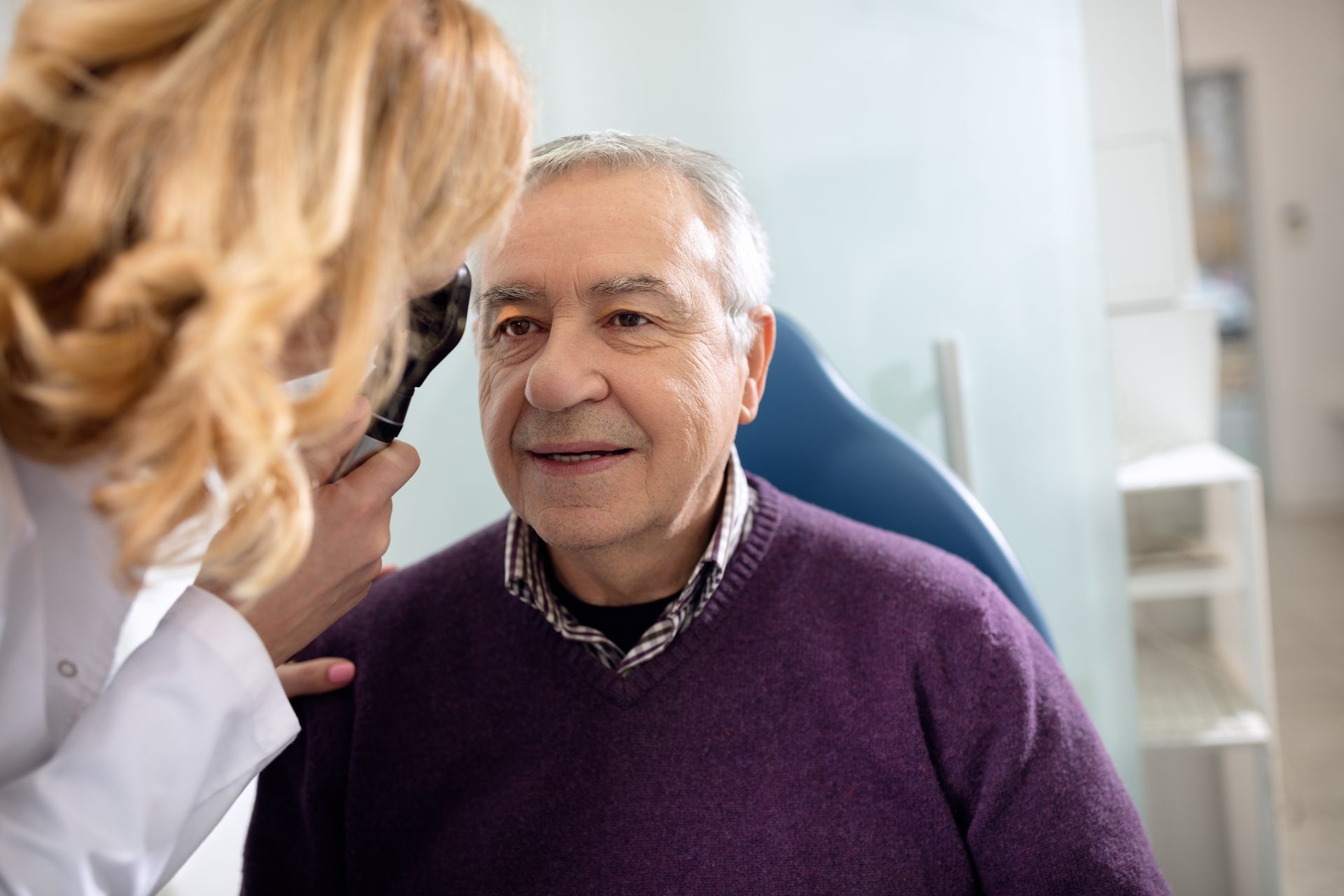 A professional wearing a white coat uses an ophthalmoscope to examine a patient’s eye in a clinical office setting.