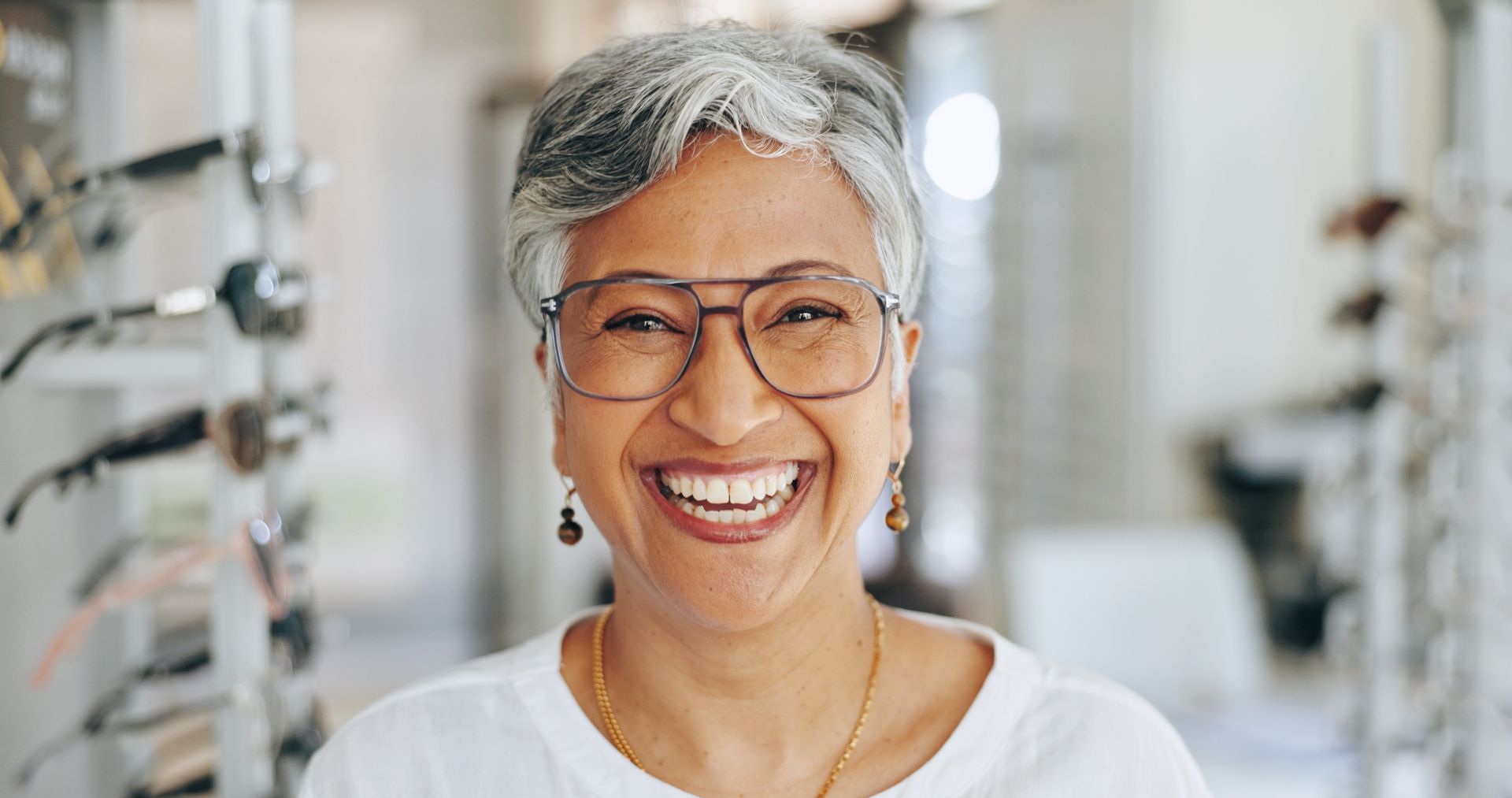 A smiling person with grey hair wears thin-rimmed glasses in an optometry shop with eyewear displays in the background.