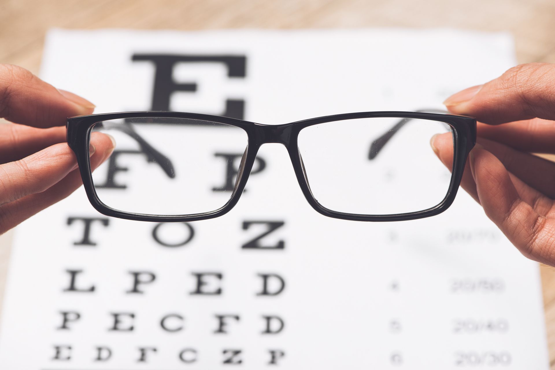 Hands hold black-rimmed glasses in front of a blurry eye chart, focusing the view through the lenses. Hands hold black-rimmed glasses in front of a blurry eye chart, focusing the view through the lenses.