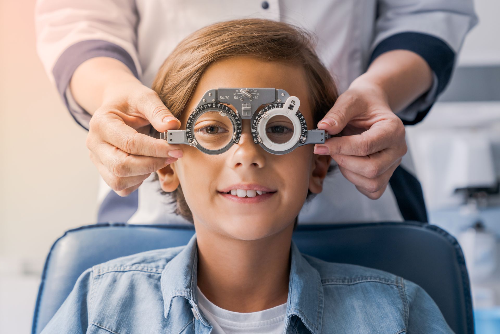 A boy wearing eyesight correction glasses & an optometrist choosing lens frames behind him.
