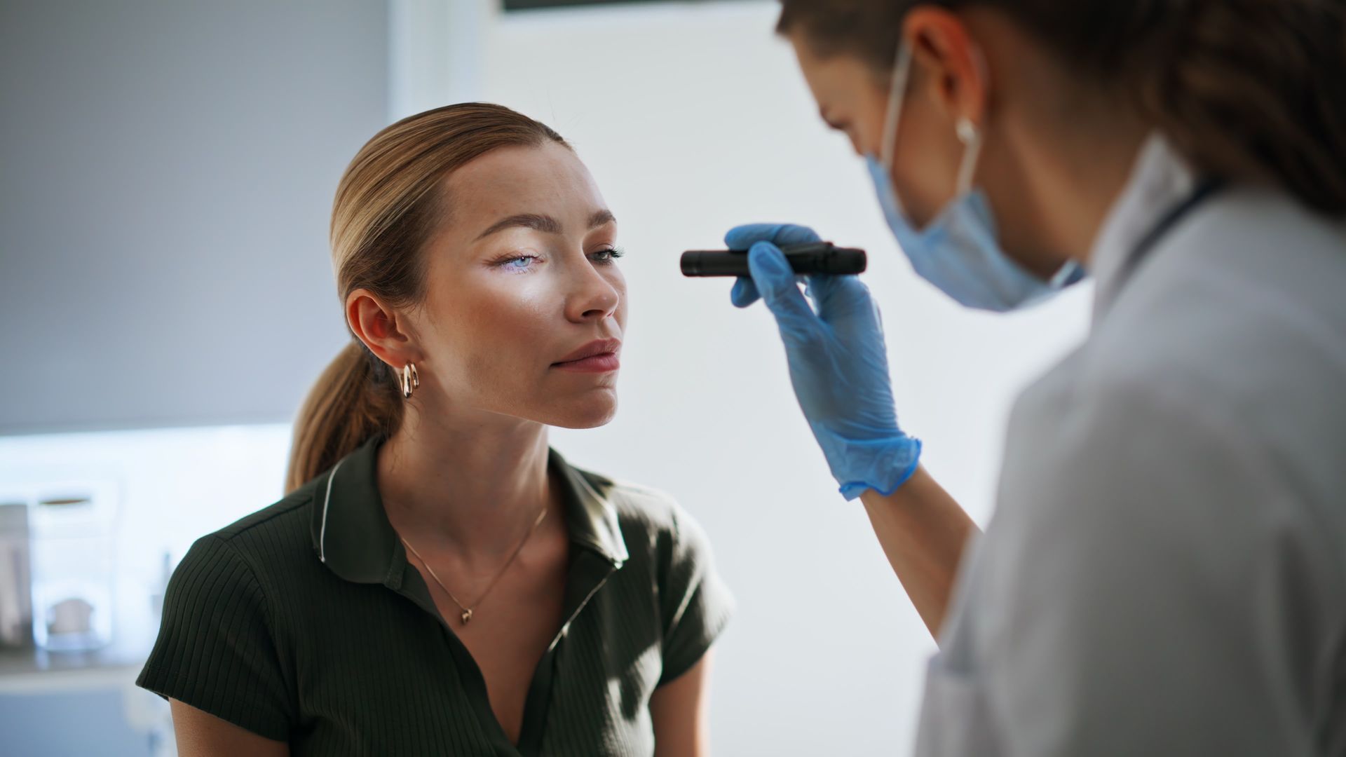 A healthcare professional wearing a mask and gloves examines a patient's eyes using a small penlight in a clinic.