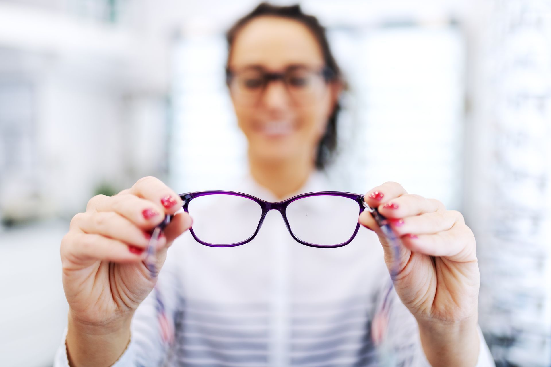 A person wearing glasses holds a pair of dark-rimmed prescription glasses toward the camera in an optical store. A person wearing glasses holds a pair of dark-rimmed prescription glasses toward the camera in an optical store.