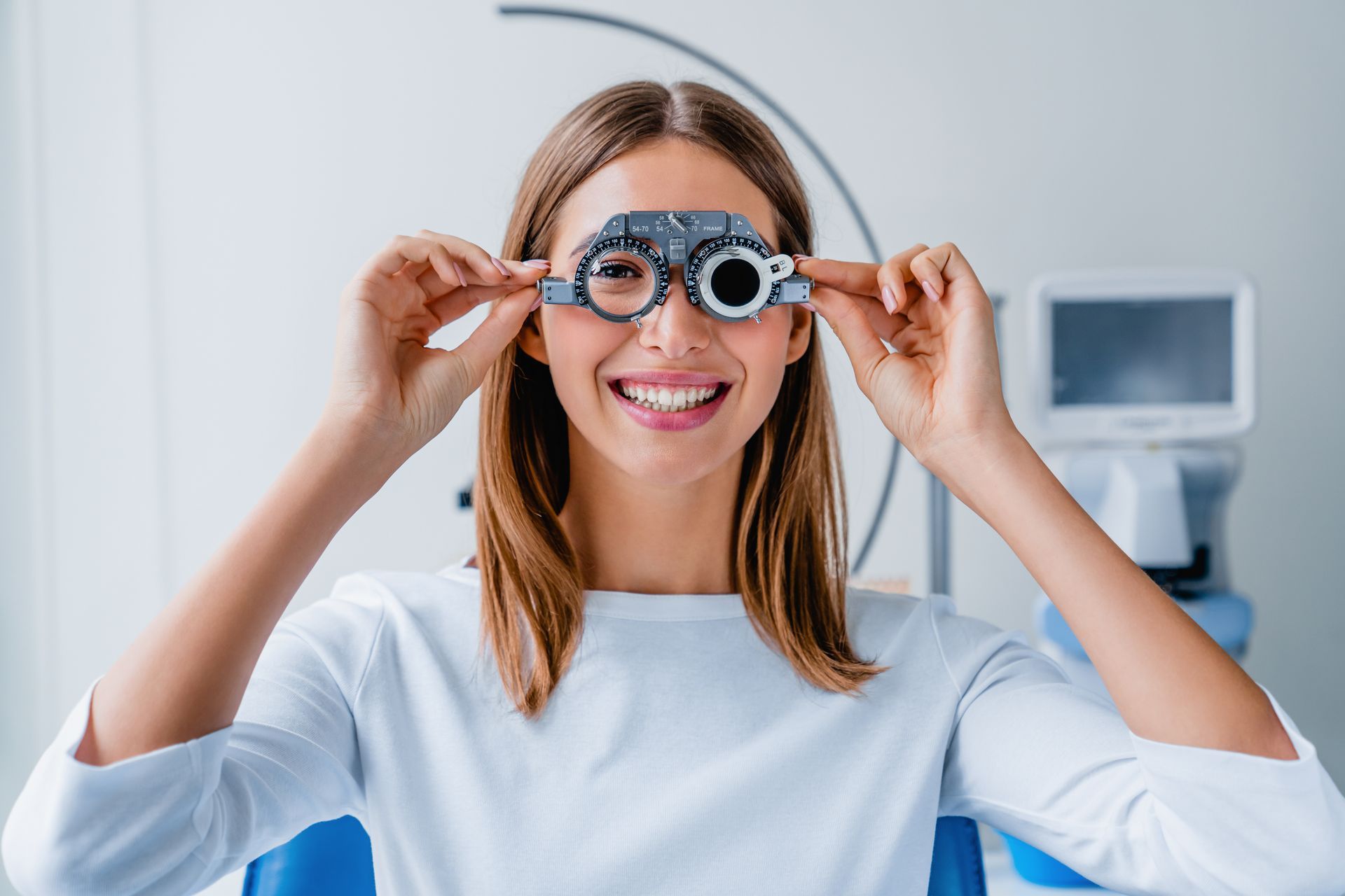 A person smiling while holding a trial frame lens set over their eyes in an eye exam setting. A person smiling while holding a trial frame lens set over their eyes in an eye exam setting.
