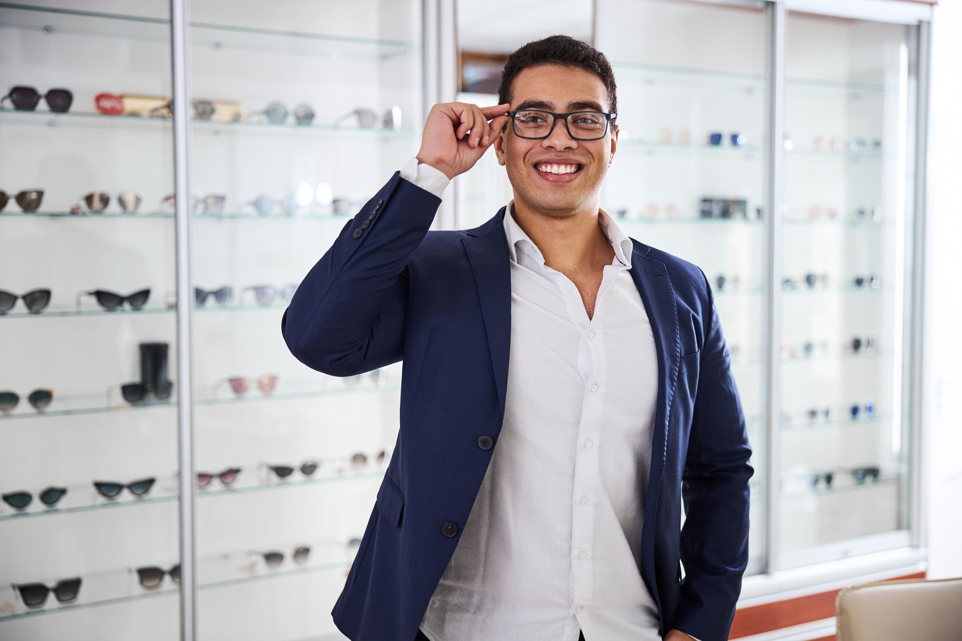 A smiling person in a blue blazer and white shirt adjusting glasses, standing in an optical store with shelves of eyewear.