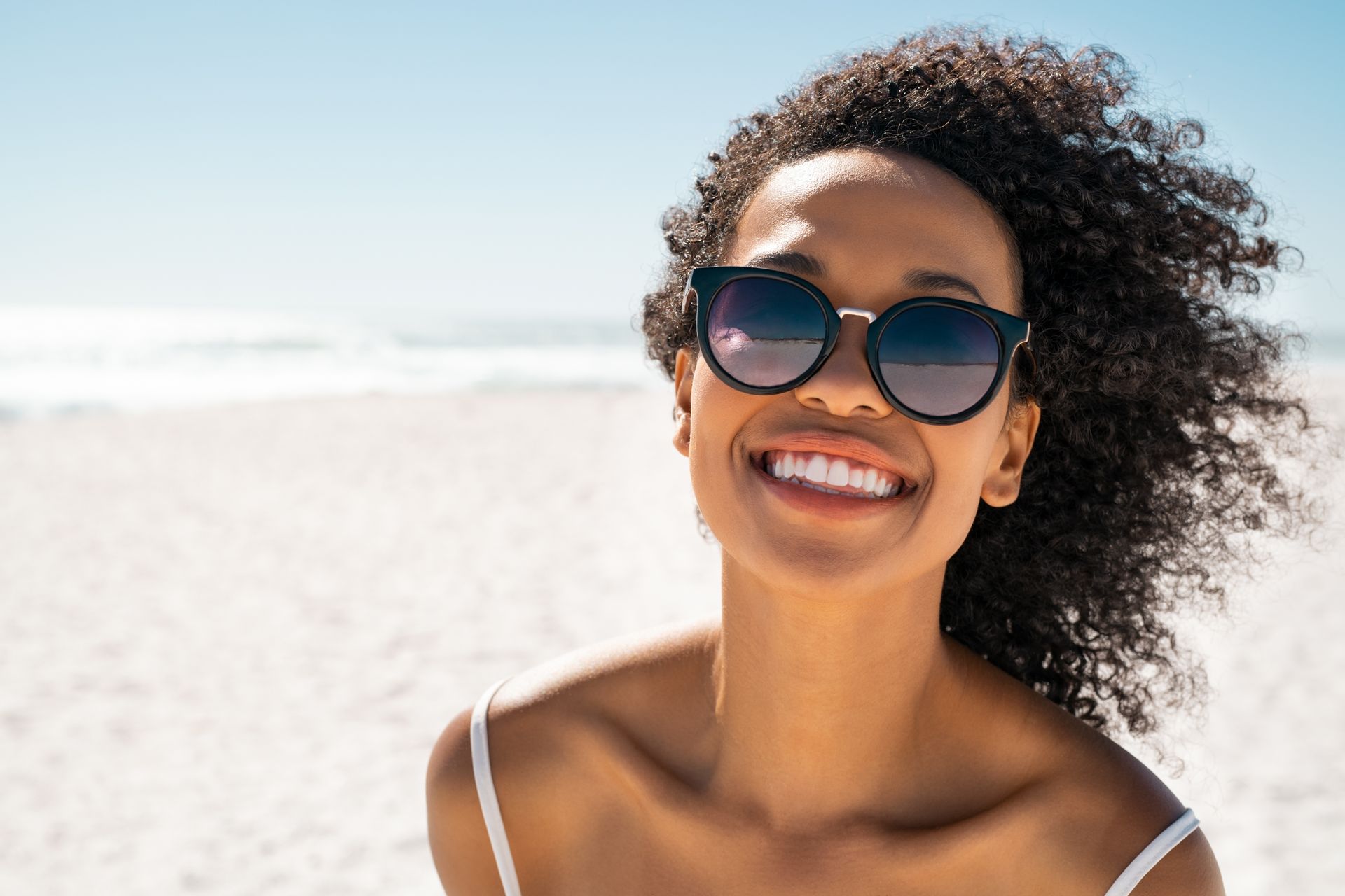 A smiling person wearing sunglasses on a sunny, white sand beach.