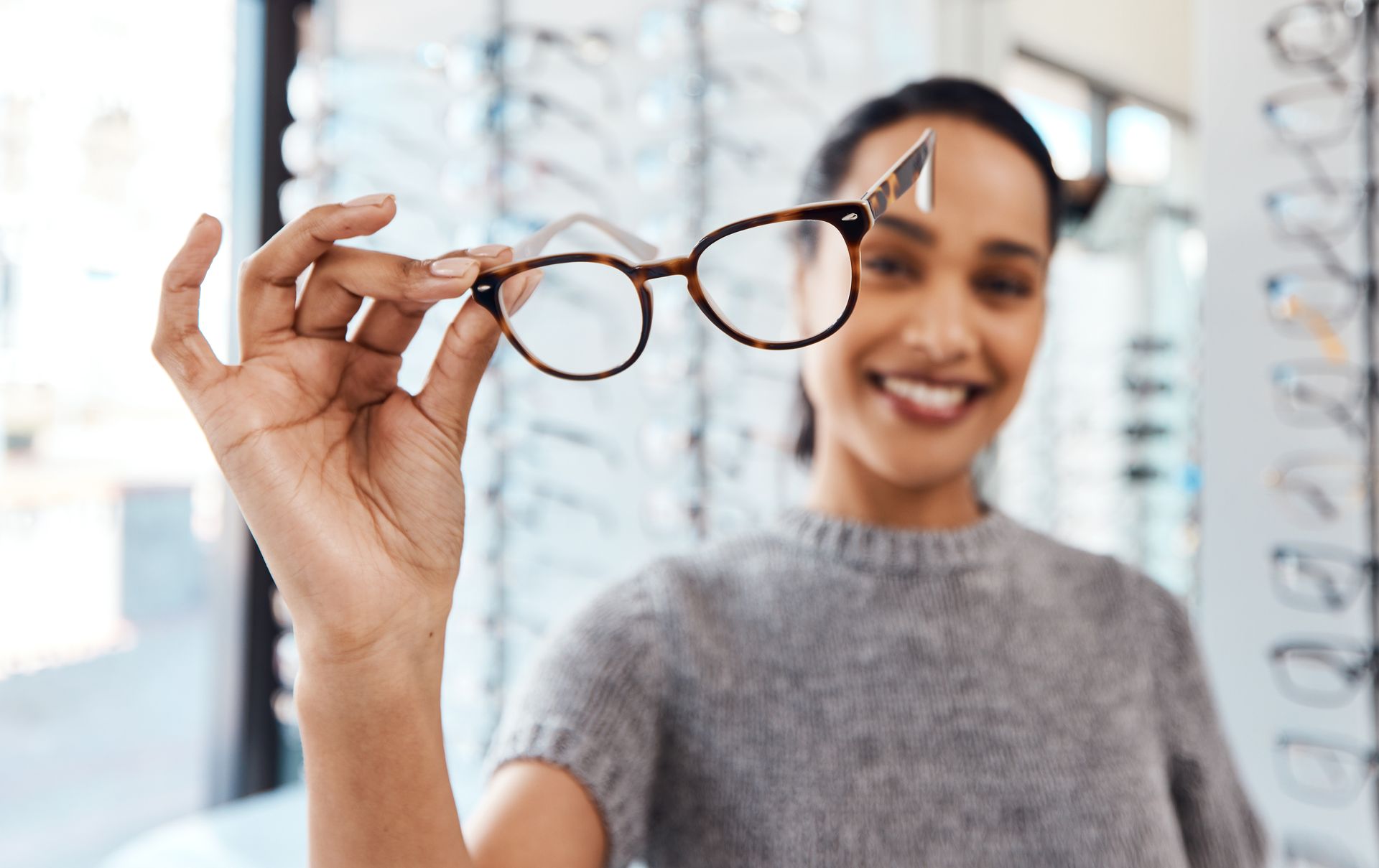 A person smiling and holding a pair of tortoise-shell glasses in front of their face inside an optical shop.