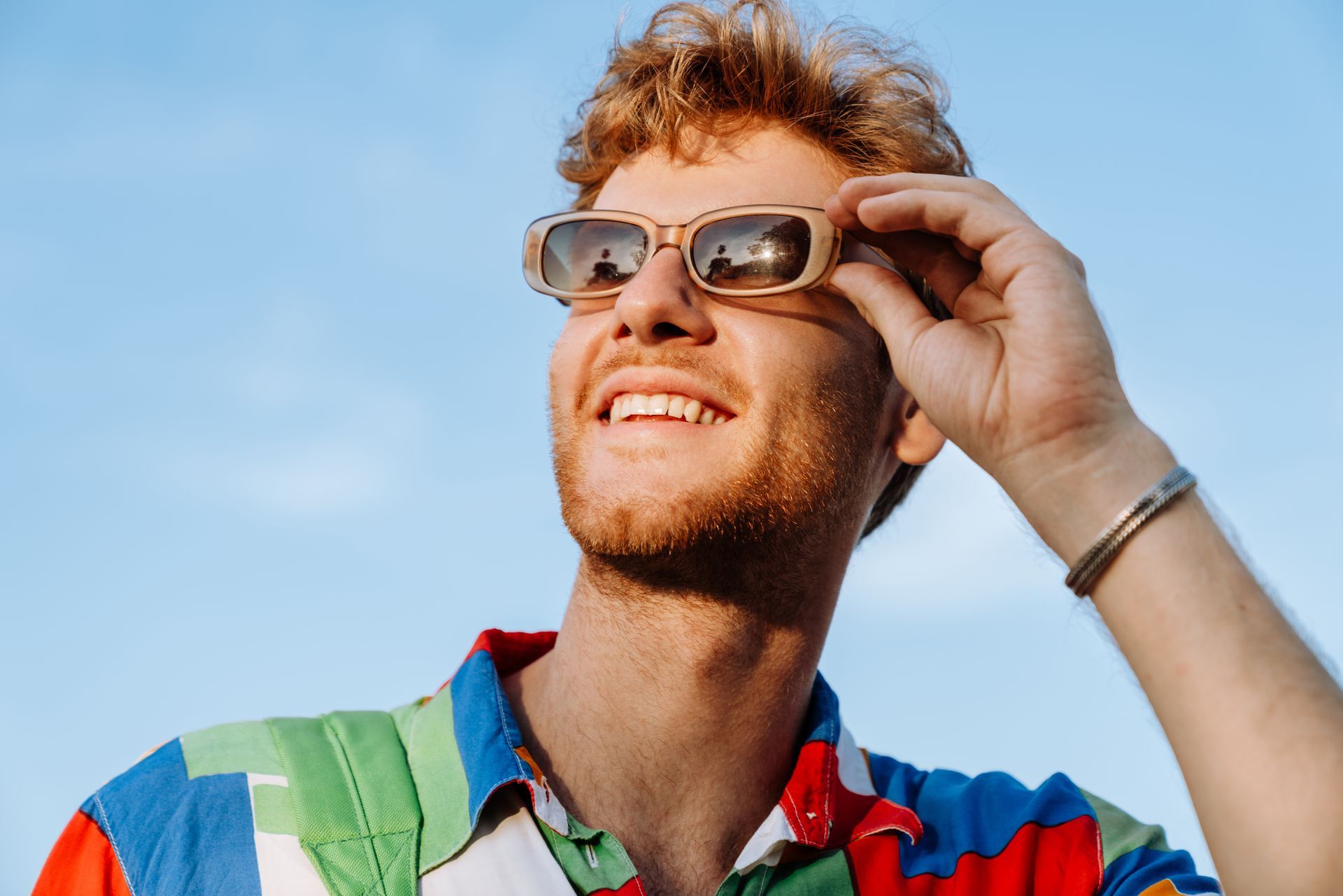 A person with light brown hair smiles while holding up amber-tinted sunglasses against a bright blue sky.