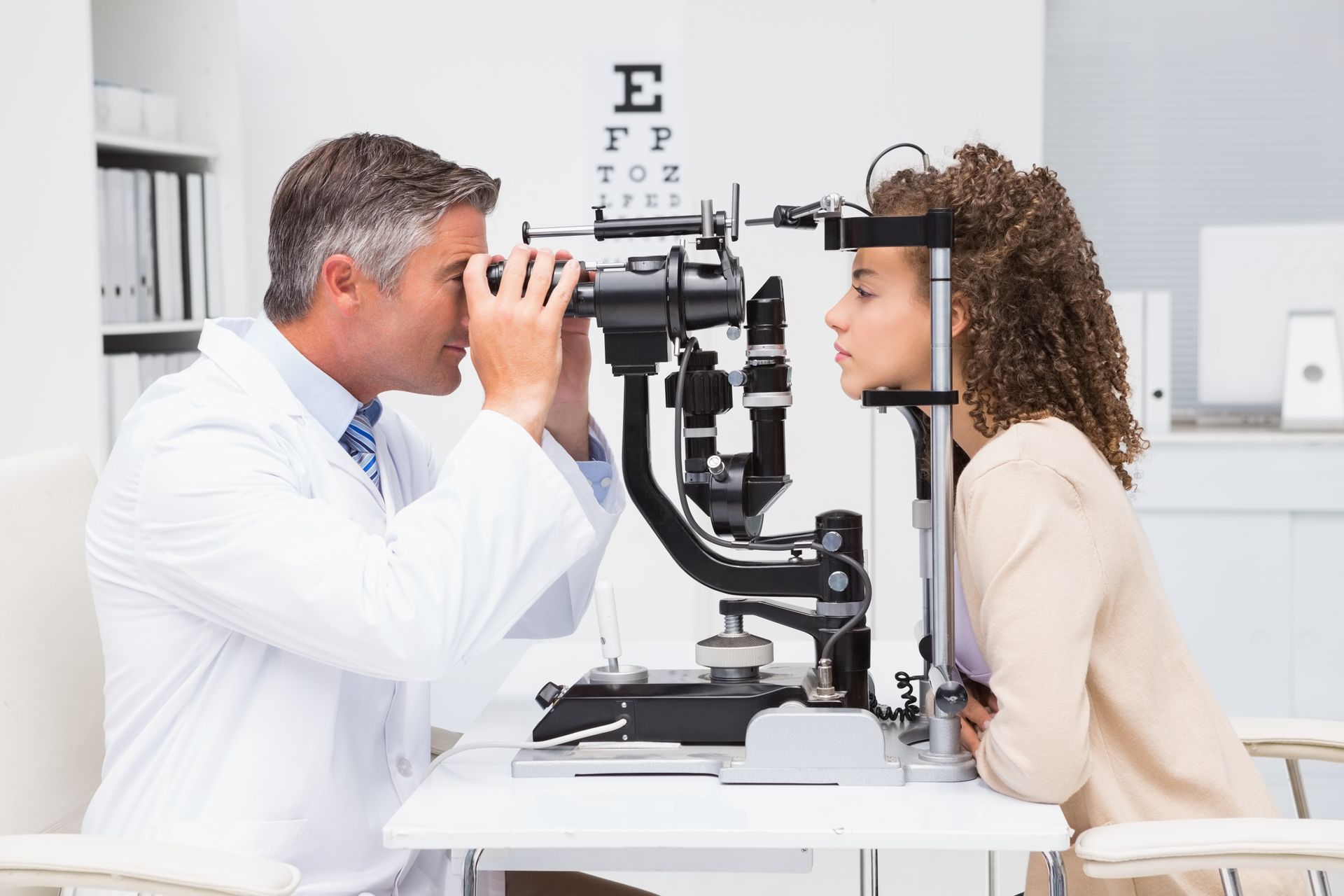 An optometrist performs an eye examination on a patient using a slit lamp in a clinical office.