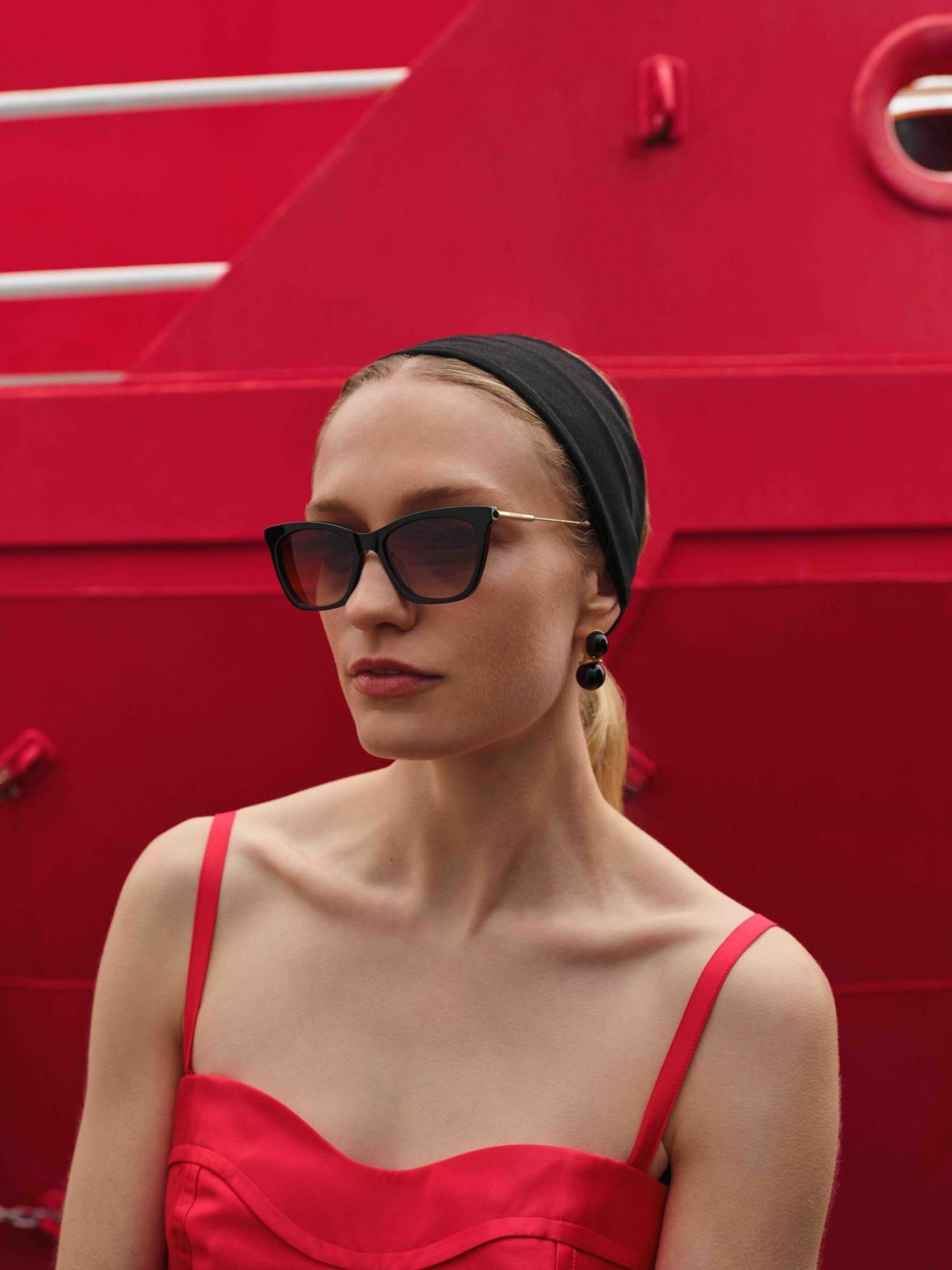 A person with light brown hair smiles while holding up amber-tinted sunglasses against a bright blue sky.