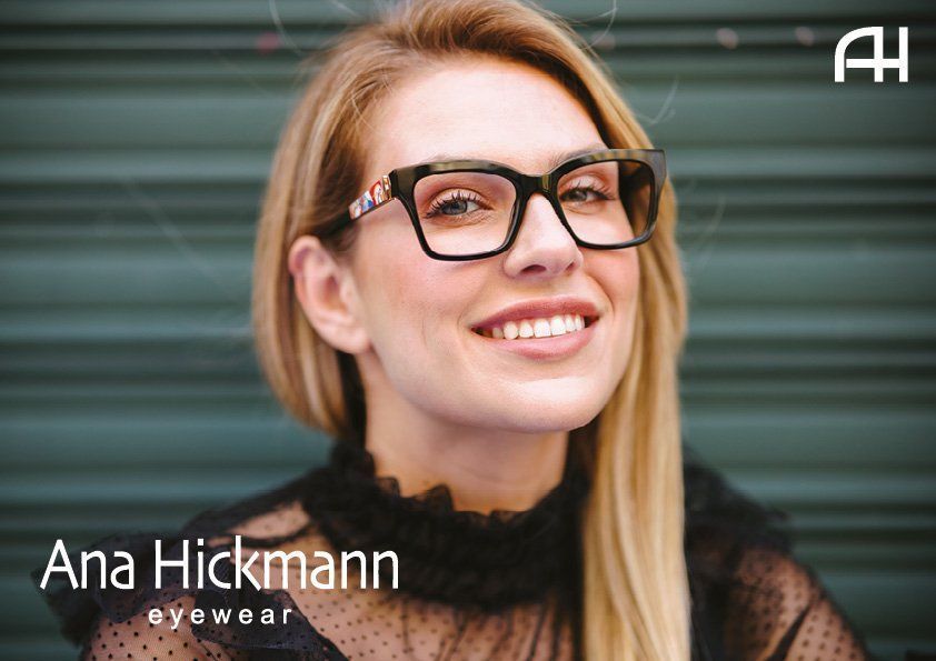 A woman with blonde hair smiling while wearing black-rimmed Ana Hickmann glasses against a dark slatted background.