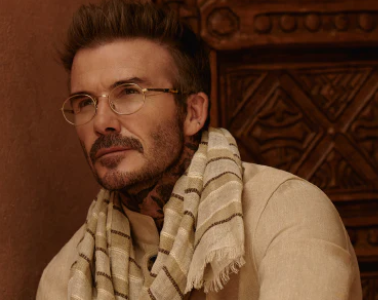 David Beckham wearing a cream striped scarf and clear-frame glasses, captured in a warm-toned, indoor portrait.