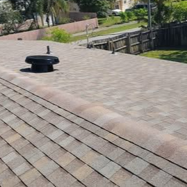 A high-angle view of a residential shingled roof with a black circular attic vent and several roof pipe stacks.