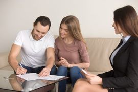 Three people discussing paperwork at a table, with one man signing a document.
