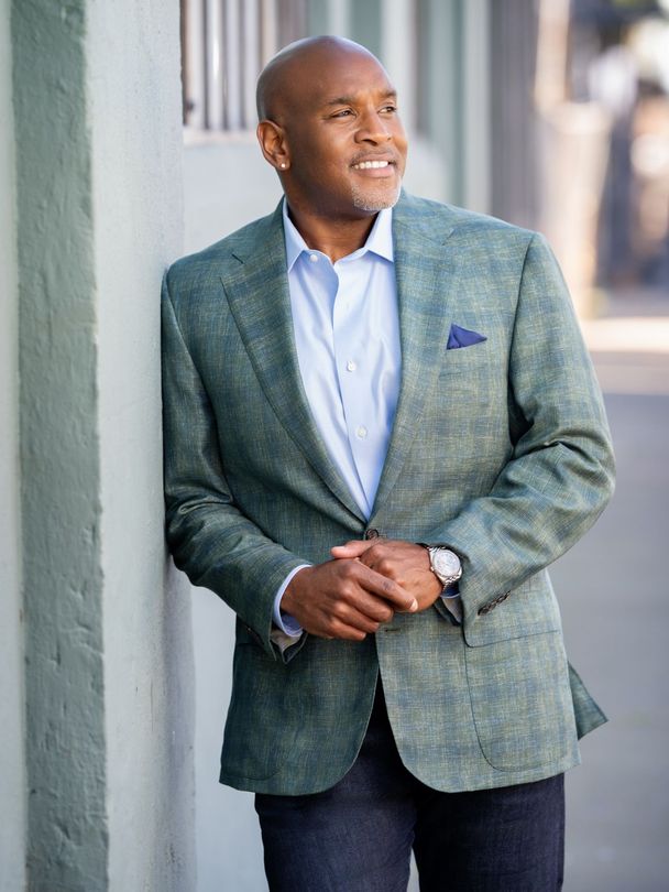 Man in a green blazer leaning against a wall, looking off to the side with a slight smile.
