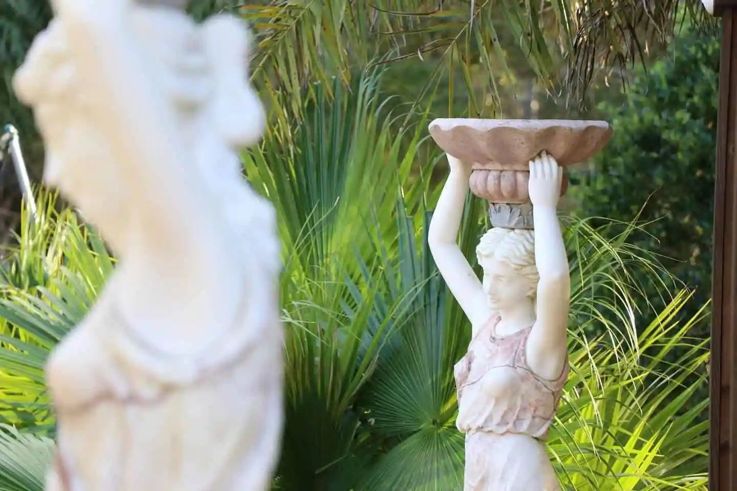 Statue of a woman holding a decorative bowl atop her head, in a garden. Blurred figure in the foreground.